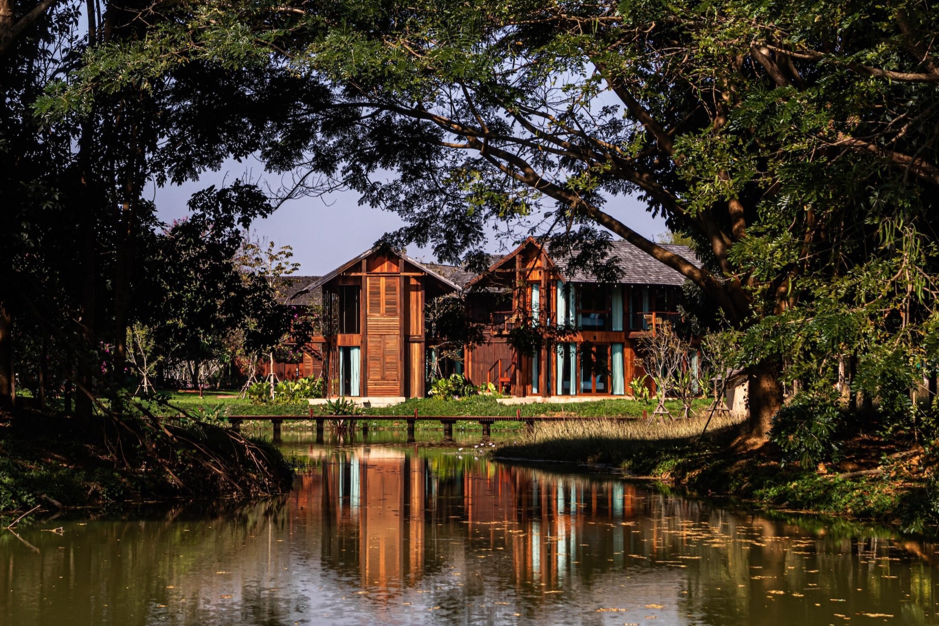 Timber cabins with gabled roofs seen across a still pond, their reflections broken by drooping tree branches in warm afternoon light