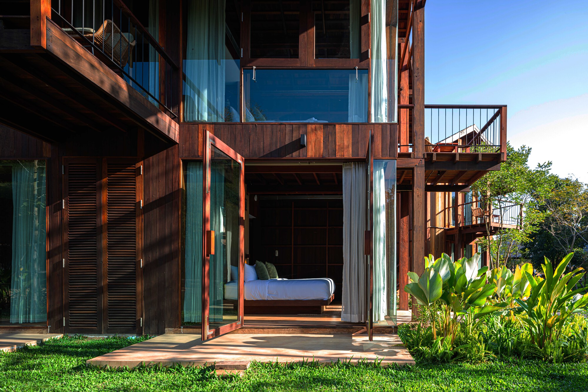 Open glazed door of a ground-floor bedroom beneath a timber balcony, with a concrete terrace and broad-leaved plants in bright sun