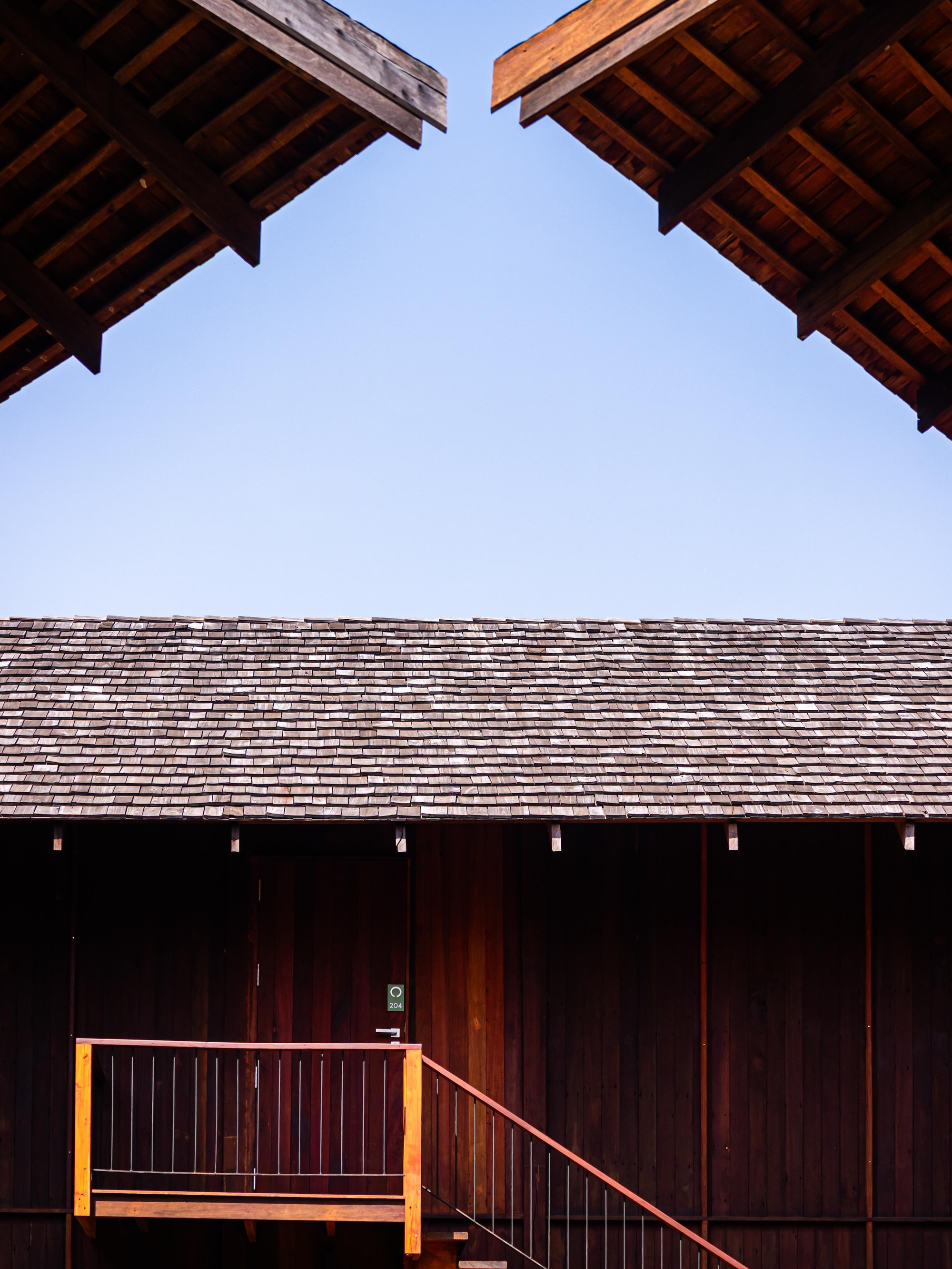 Exposed rafters of two roof eaves framing a shingled roof and timber wall with entry stair beneath a clear sky