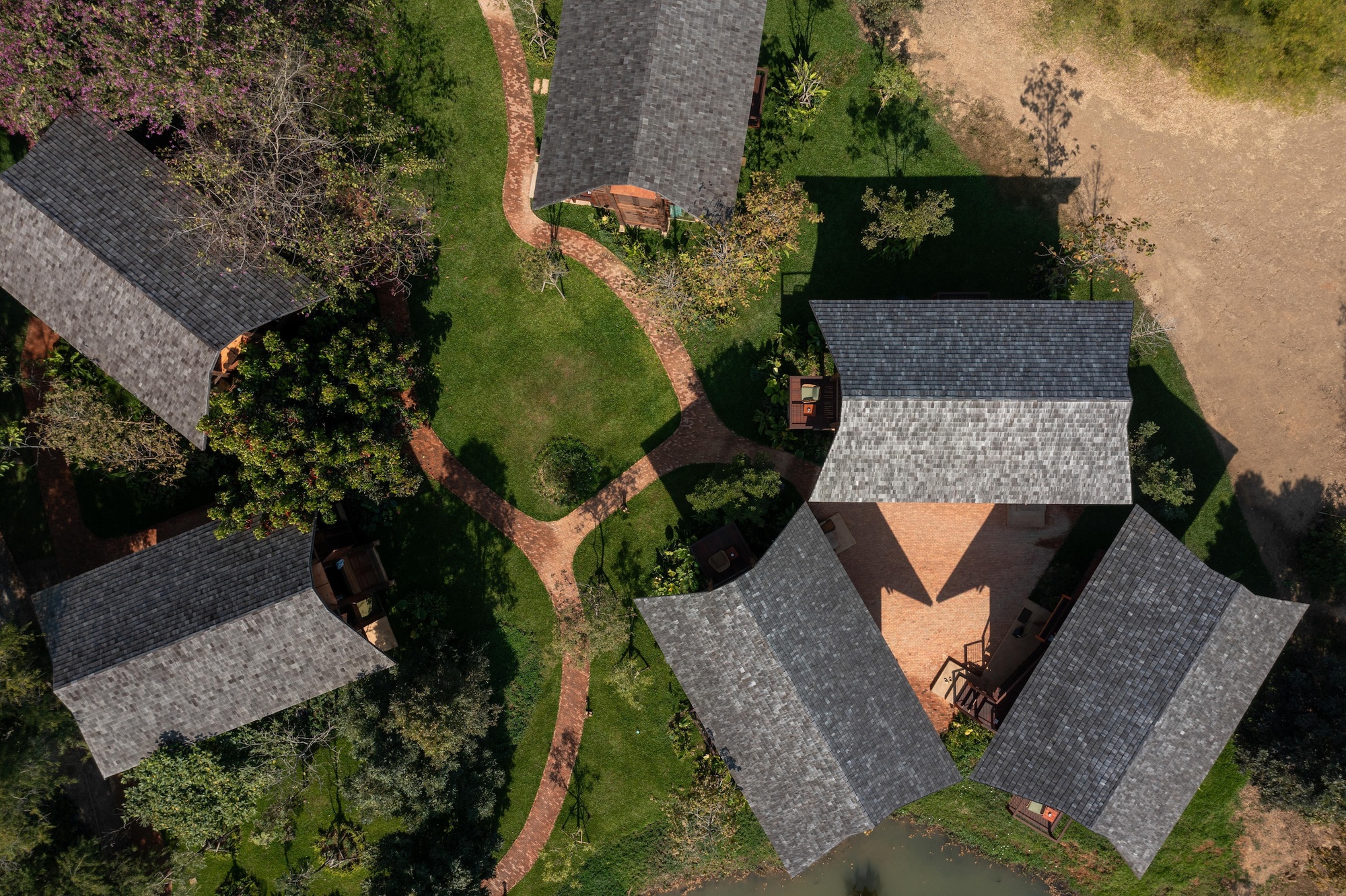 Aerial view of grey-shingled cabin roofs arranged around a brick path, lawn and small pond in afternoon light