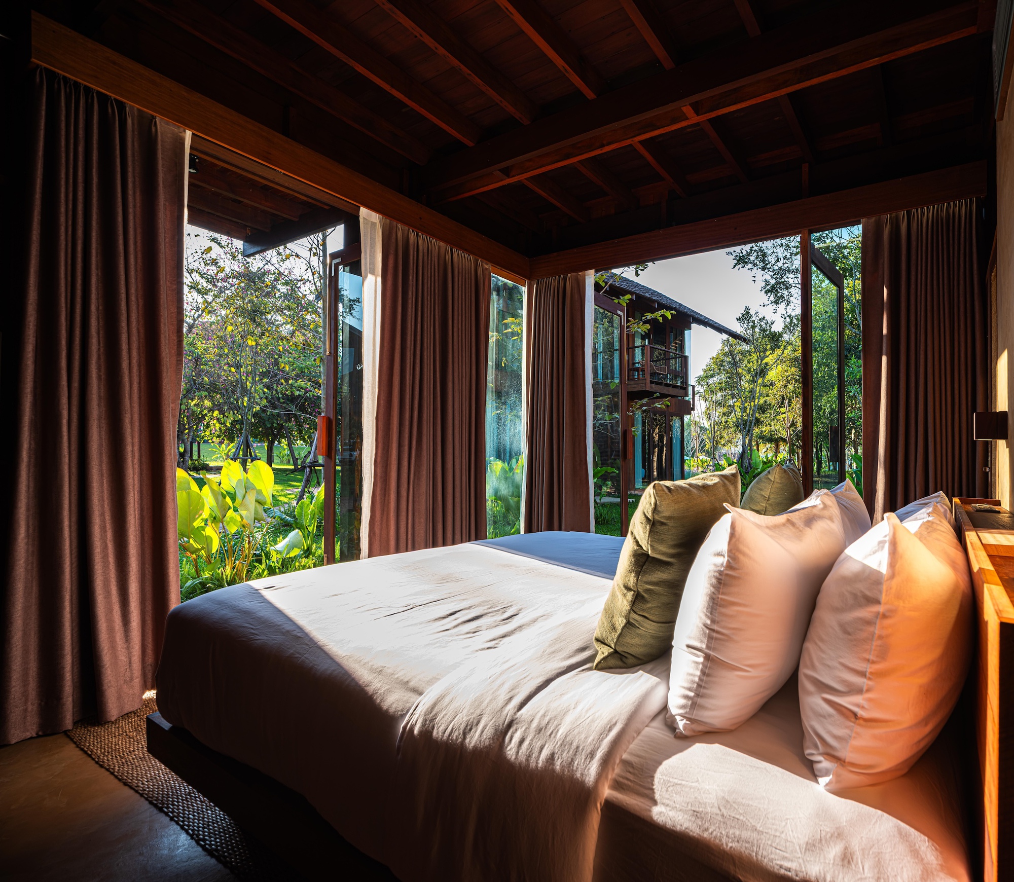 Bed with olive and white pillows beside floor-to-ceiling glass doors and brown curtains, opening onto a sunlit garden