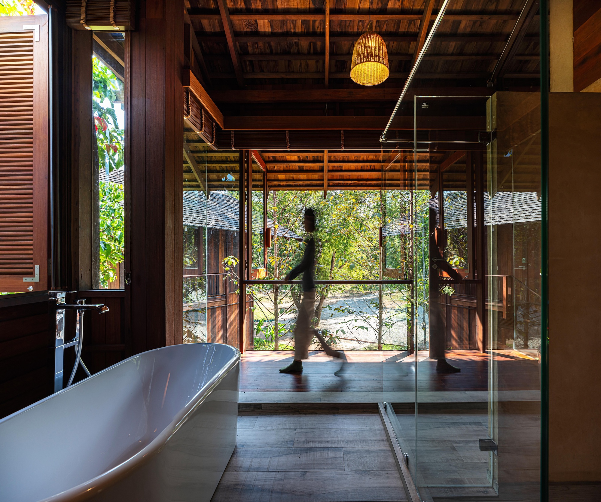 Freestanding white bathtub and glass shower enclosure facing a timber veranda, with a blurred figure walking past in daylight