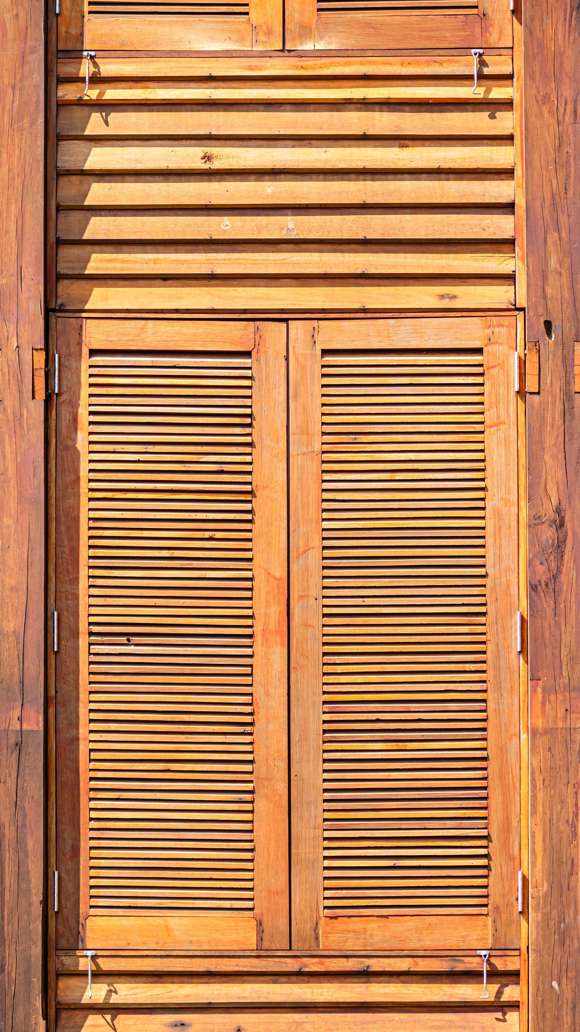 Pair of louvered timber shutters with a fixed louvered panel above, lit by strong direct sunlight