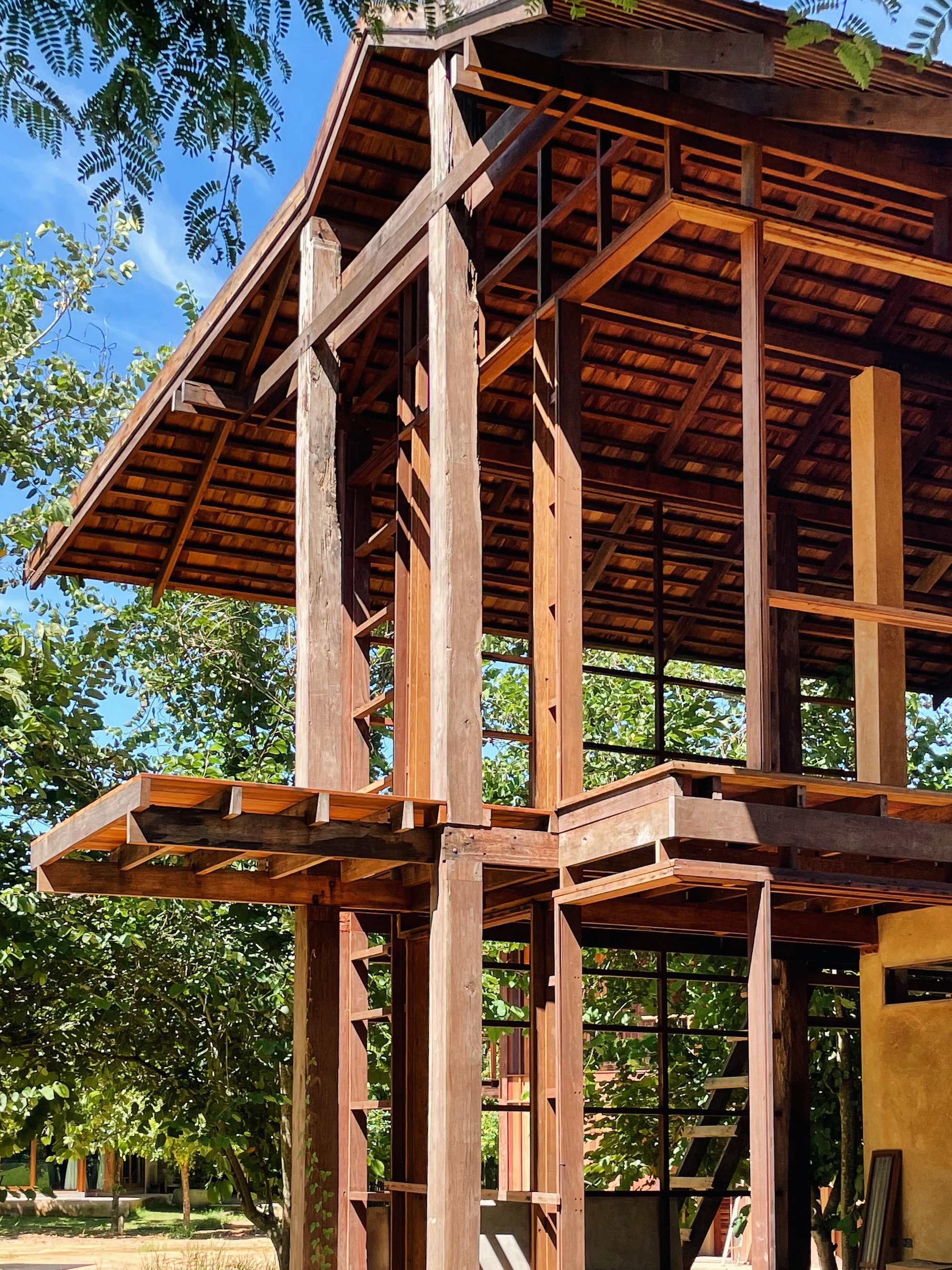 Timber frame of a two-storey structure with clay roof tiles and open floor platforms, seen through leafy trees under blue sky
