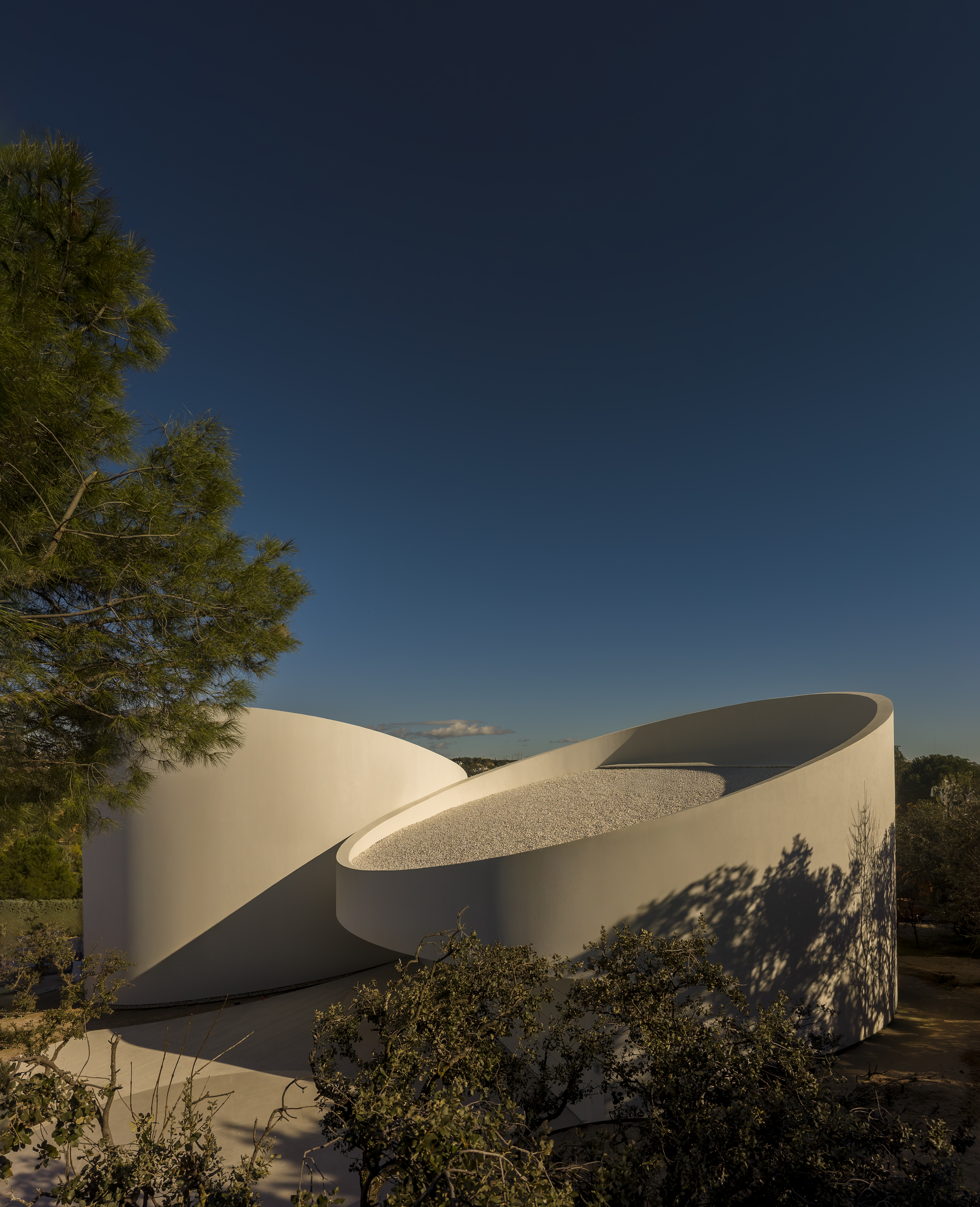 Two white cylindrical volumes with gravel-filled tops, tree shadows falling across the curved wall under a deep blue evening sky