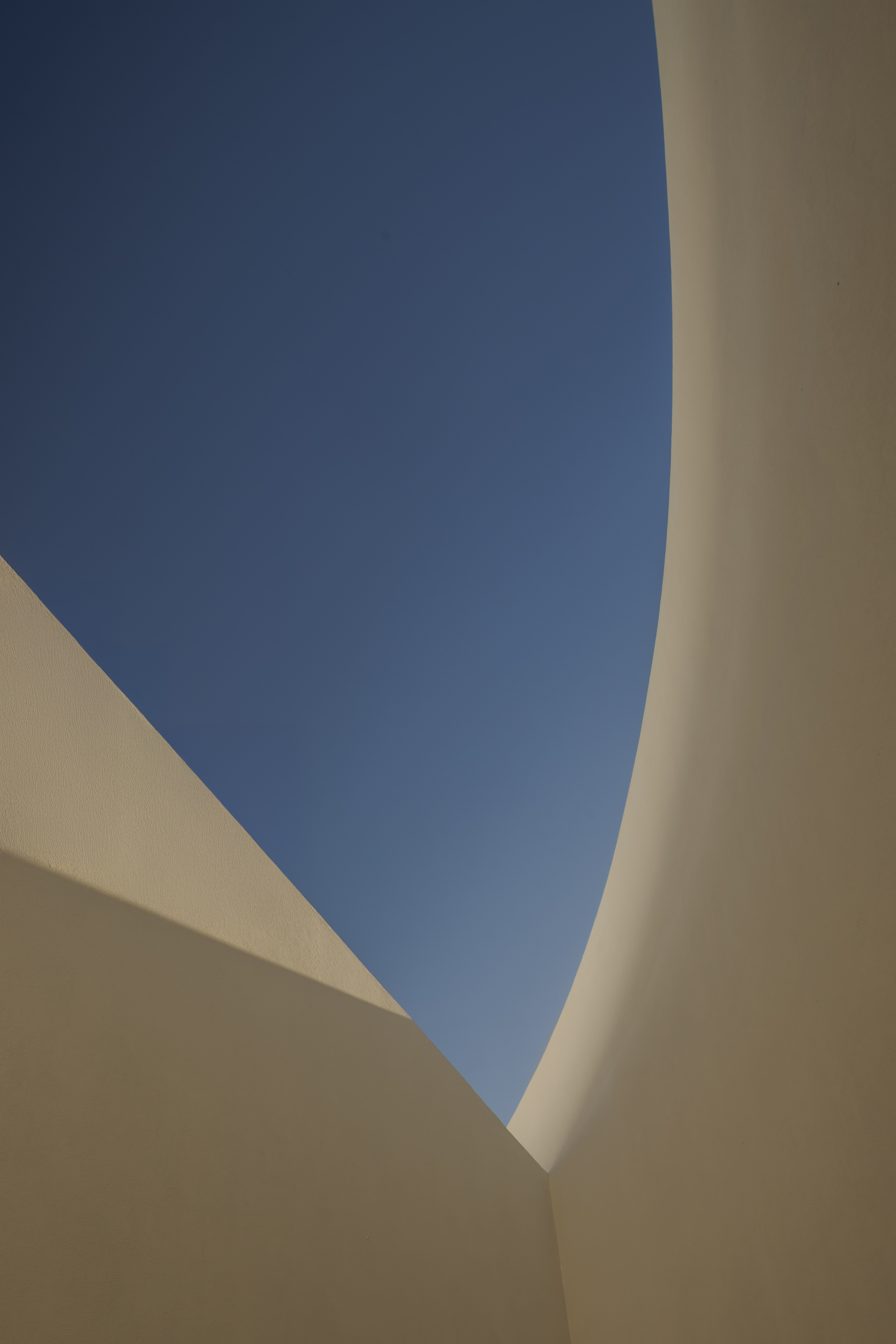 Low angle looking up between curved and angled white walls to a narrow band of blue sky