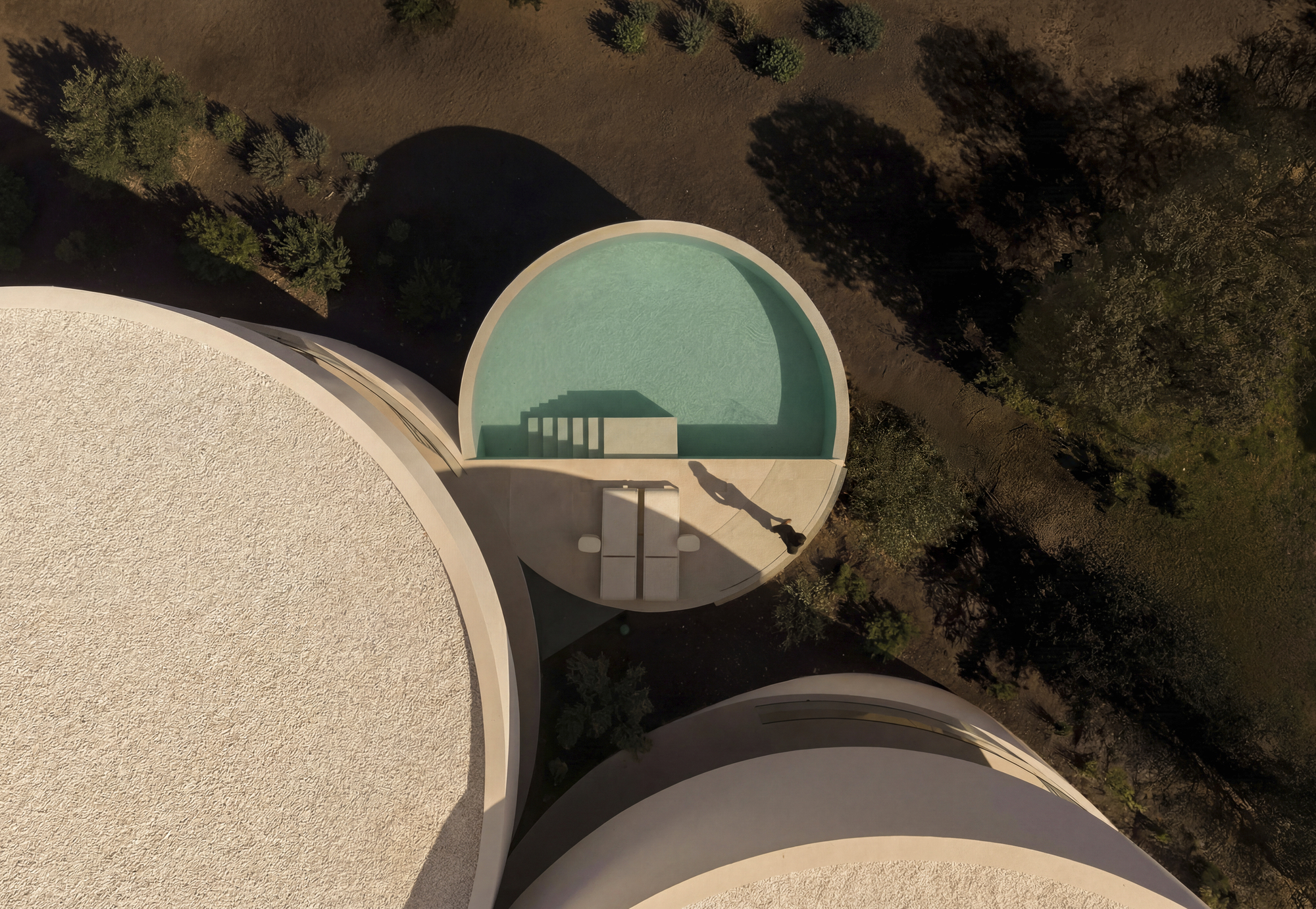 Top-down view of a semicircular pool with steps and two lounge chairs between circular roofs, a long shadow cast across the deck