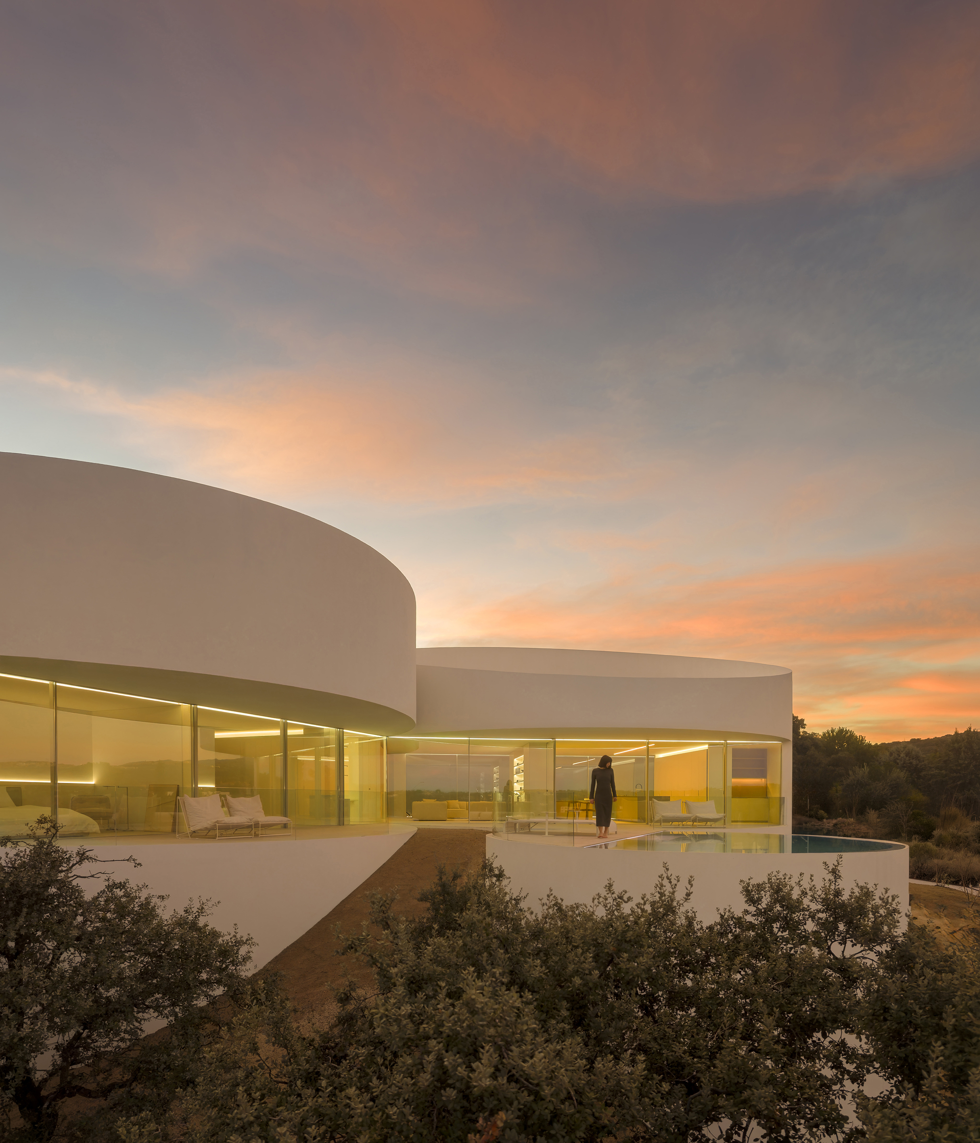 Curved white house with glazed lit living spaces and a woman standing at the terrace edge under a pink dusk sky