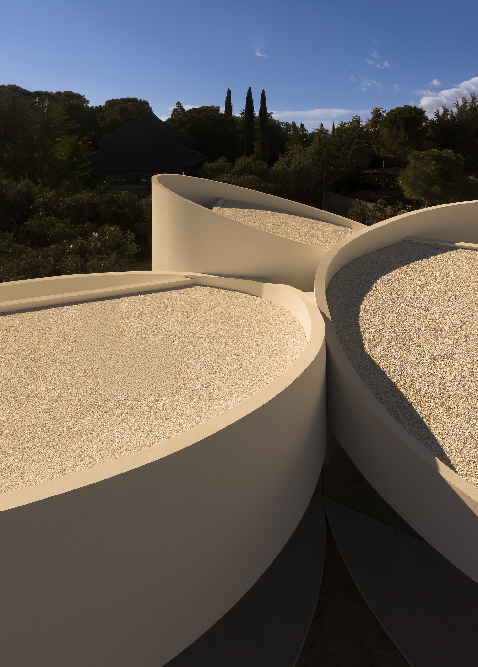 Gravel-covered circular roof terraces with curved white parapet walls, seen against cypress and pine trees in low afternoon light