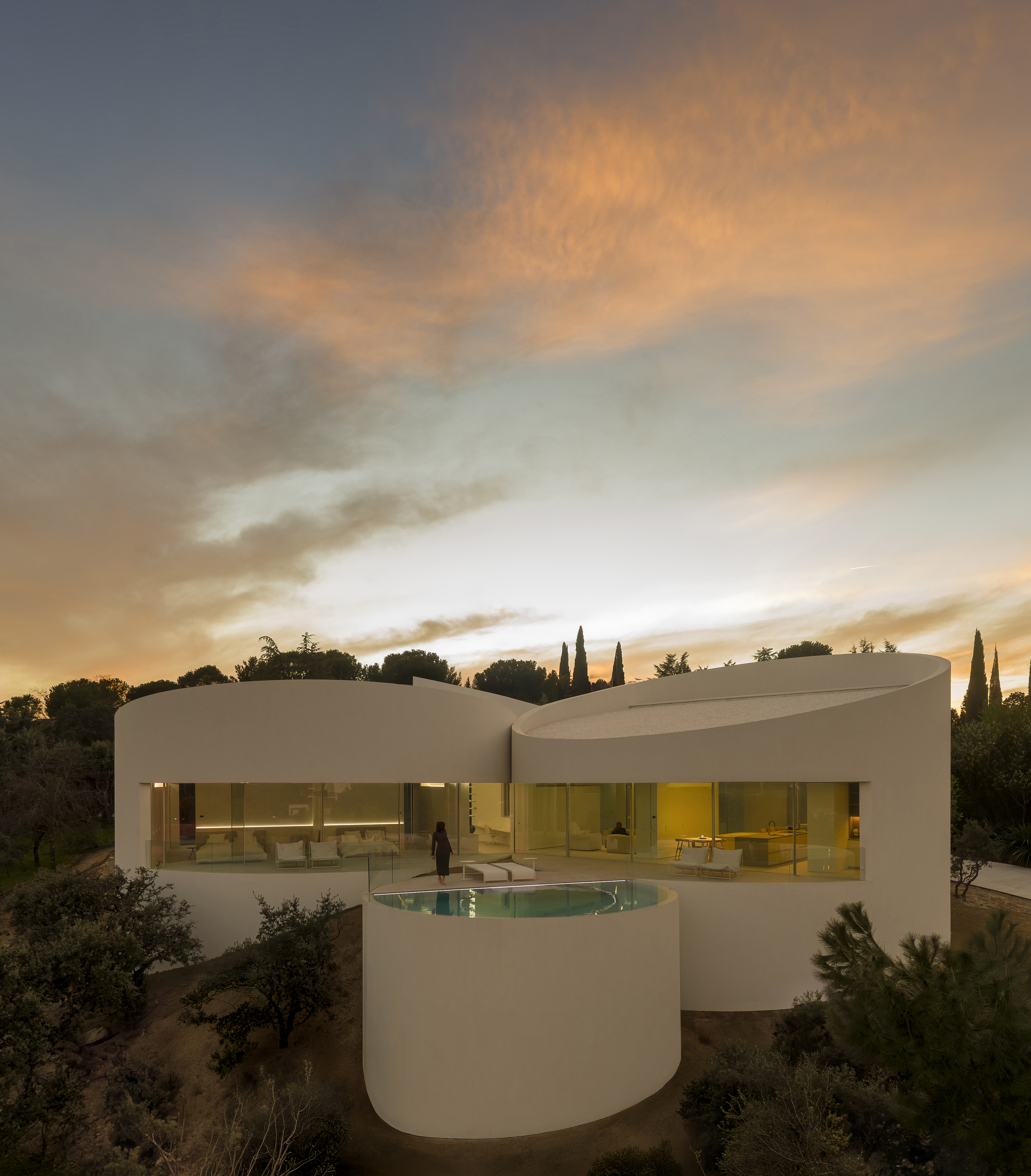 Cylindrical white volumes with a glazed living level and a round pool, a figure standing before them under a sunset sky