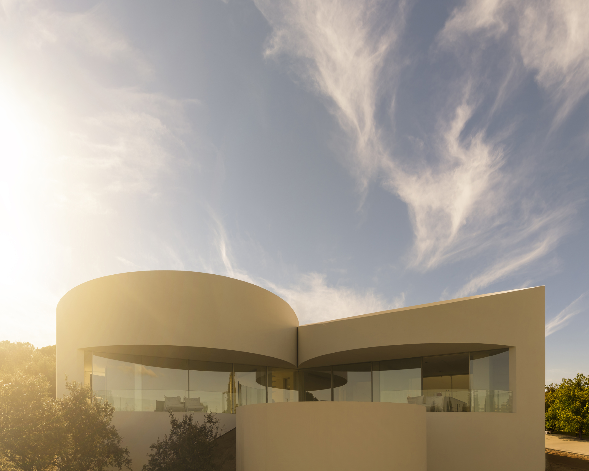 Curved white walls with a wraparound glazed band and a rounded balcony, backlit by low sun and wispy cirrus clouds