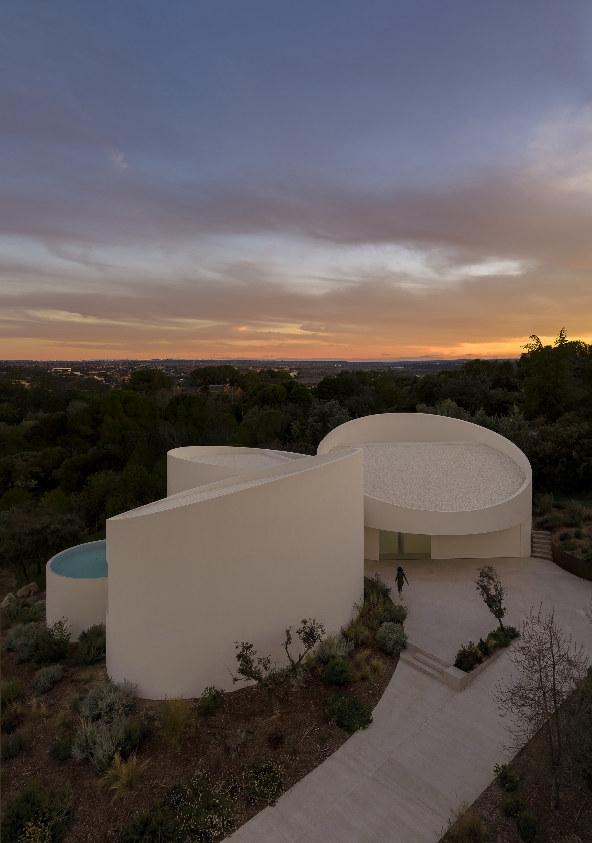 Elevated view of the curving white walls, round pool and gravel roof, with a person crossing the paved entrance at dusk