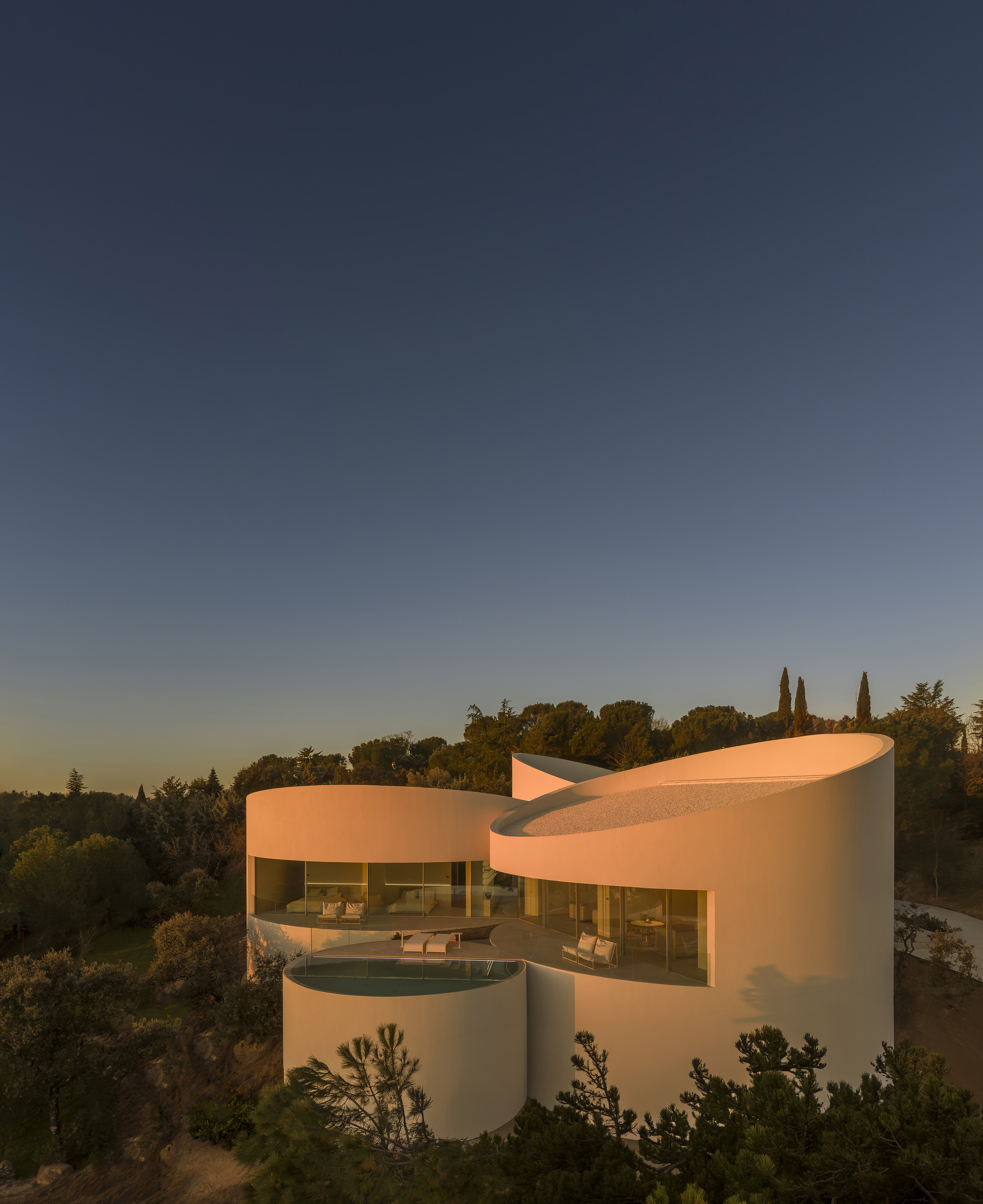 Cylindrical white volumes with a glazed terrace and small pool, lit orange by low sun above a pine and cypress treeline