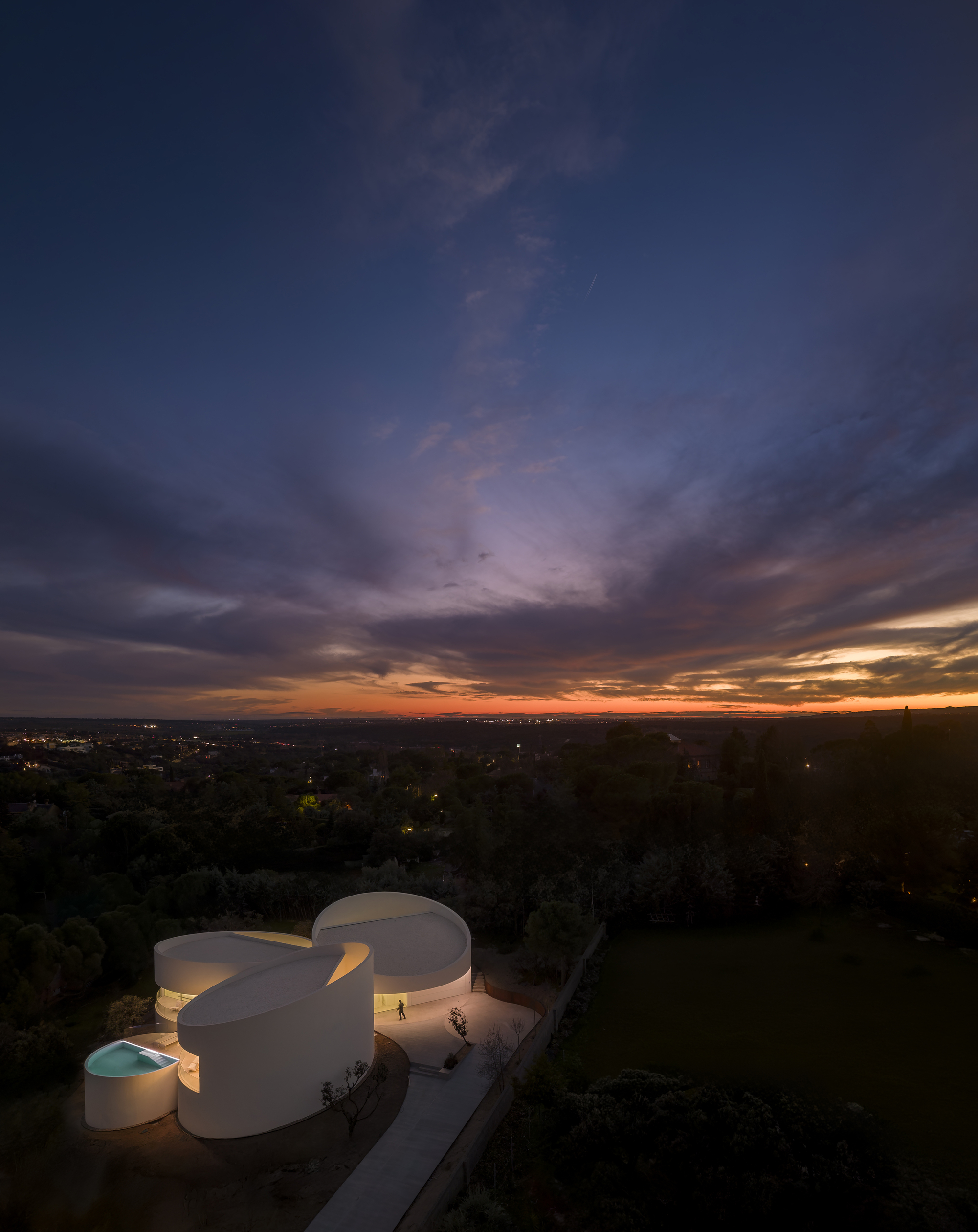 Aerial view of the white circular volumes glowing at dusk, with a lone figure by the entrance under a streaked purple sky
