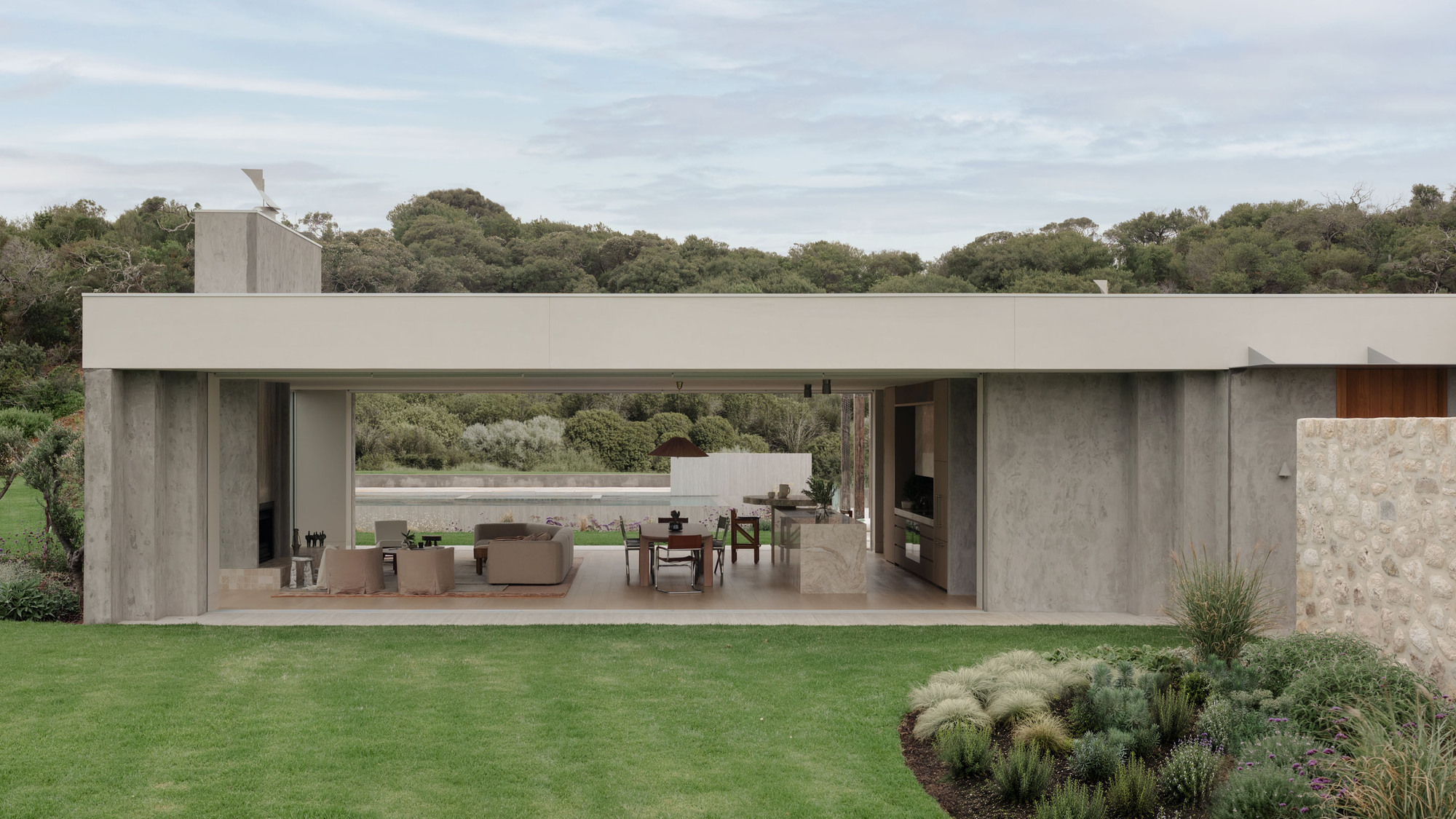 Open-sided concrete pavilion with living and dining area framed by a flat roof, lawn and grass bed under soft cloud