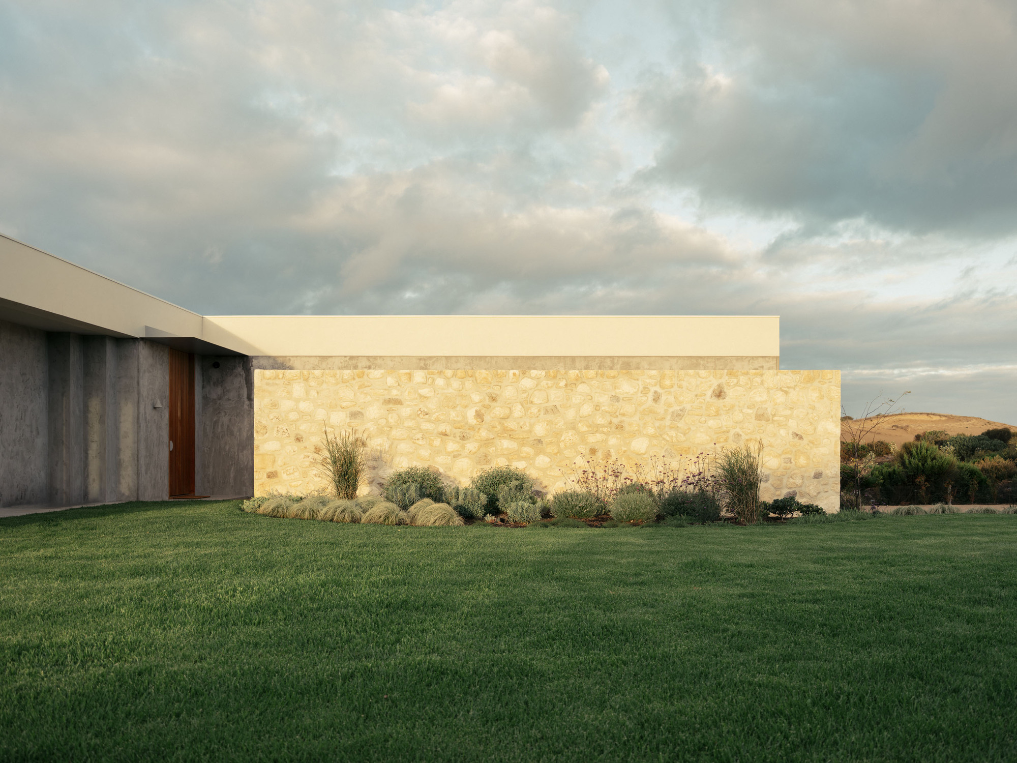 Rubble stone wall and timber door beside board-marked concrete, lit by warm low sun over a lawn and grass planting