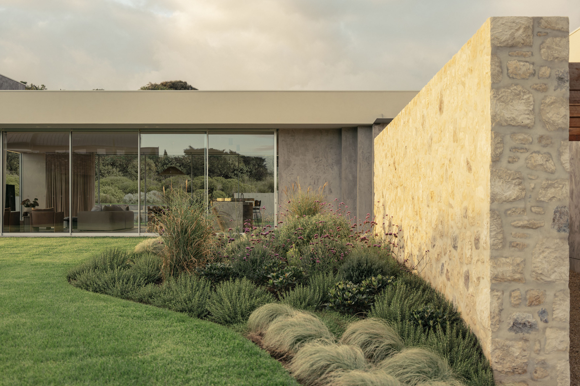 Glazed living room corner beside a tall rubble stone wall, with curved planting of grasses and verbena in overcast light