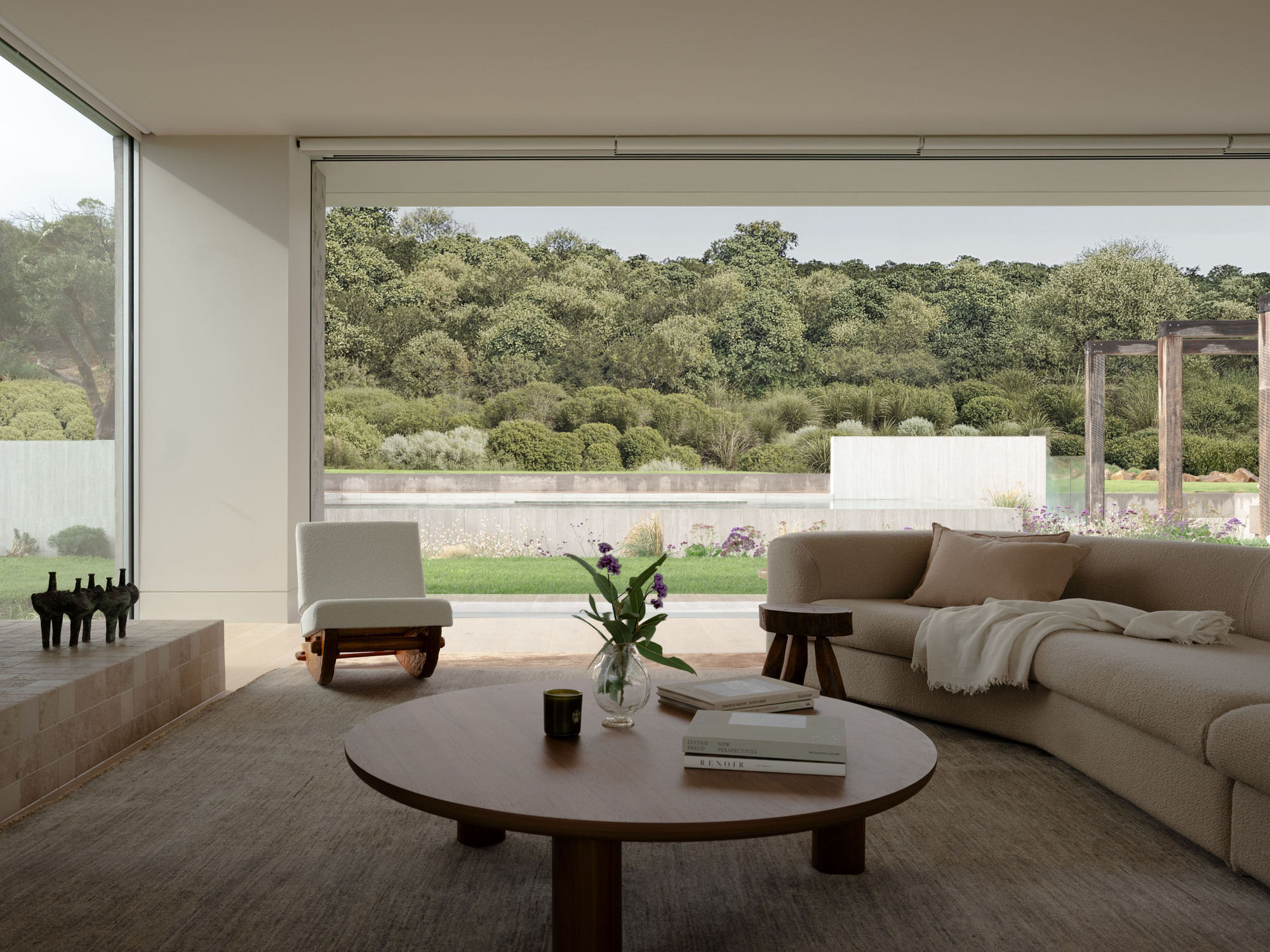 Living room with curved boucle sofa, round timber coffee table and rocking chair opening through glass to a hedged landscape
