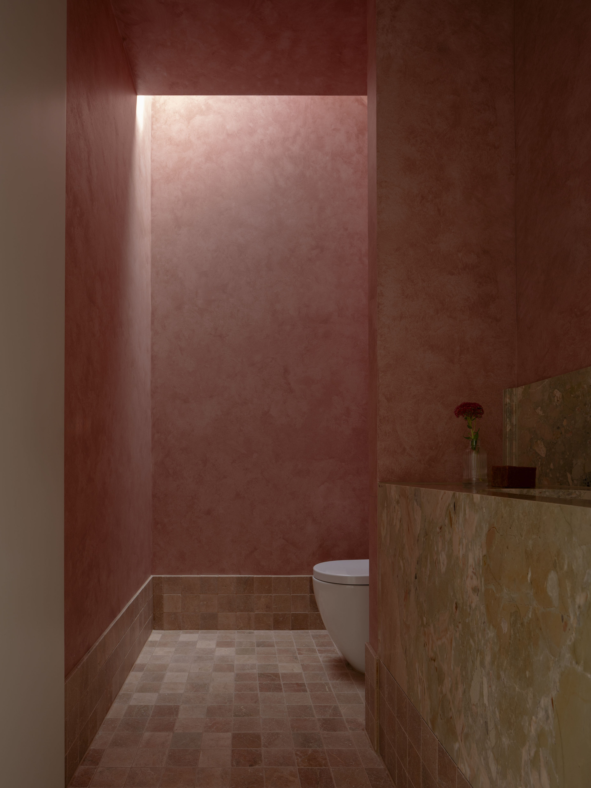 Narrow powder room with pink plastered walls, small square floor tiles and a marble basin lit by a slot of daylight