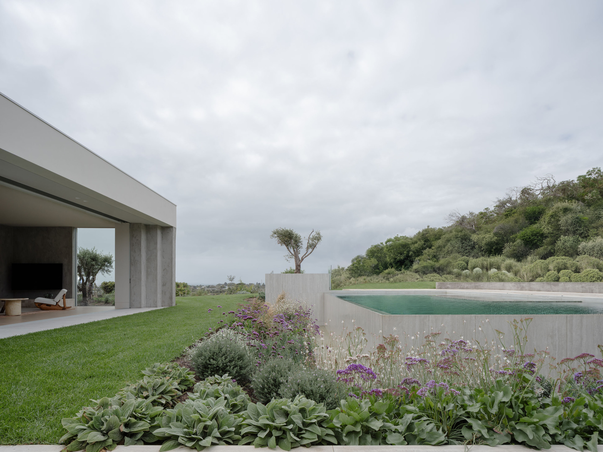 Flat-roofed pavilion with slim concrete piers beside a raised pool and perennial bed under an overcast sky