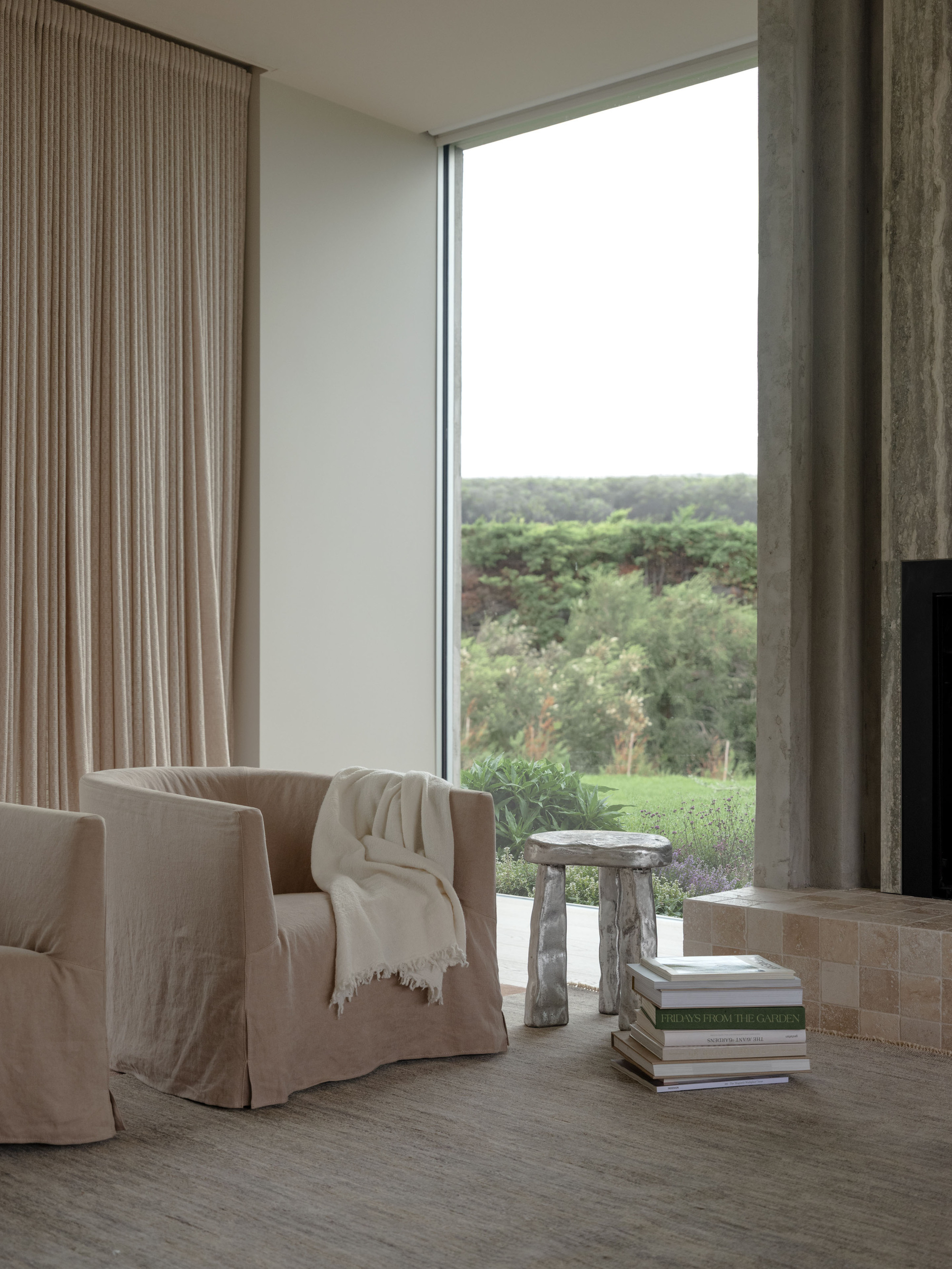 Linen slipcovered armchair and carved stool beside a corner window and travertine fireplace surround, with a green hillside outside