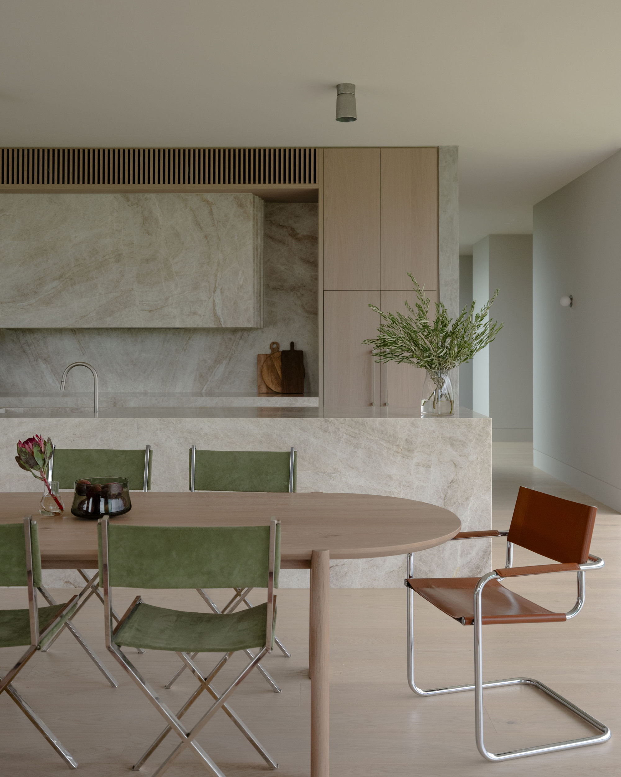 Stone-clad kitchen island and oak cabinetry behind an oval timber dining table with green folding chairs and a leather cantilever chair