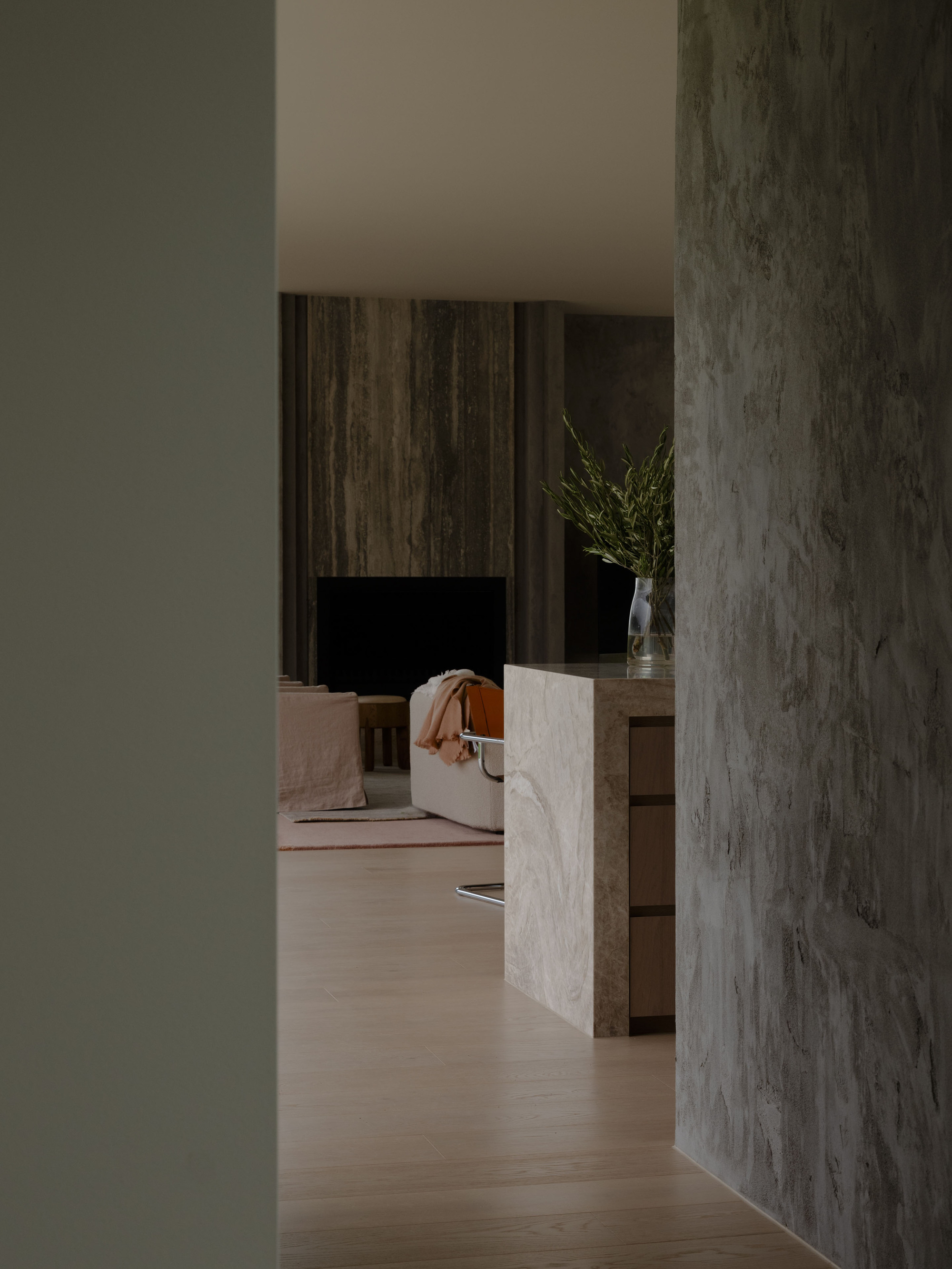 Framed view through a gap between plastered walls toward a stone island and a darker fireplace wall in low light