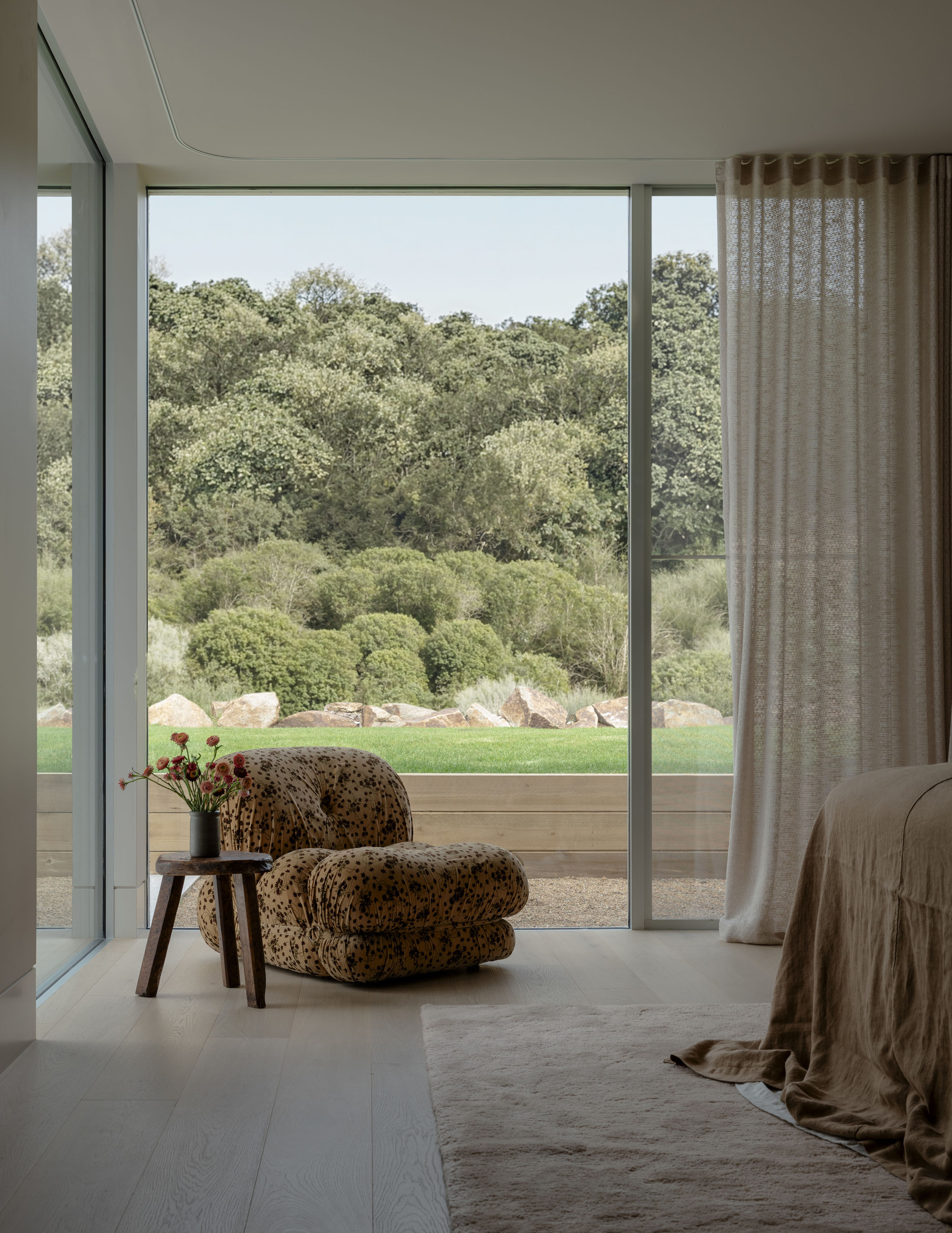 Leopard-print lounge chair and wooden stool by a floor-to-ceiling window facing boulders, lawn and a wooded slope