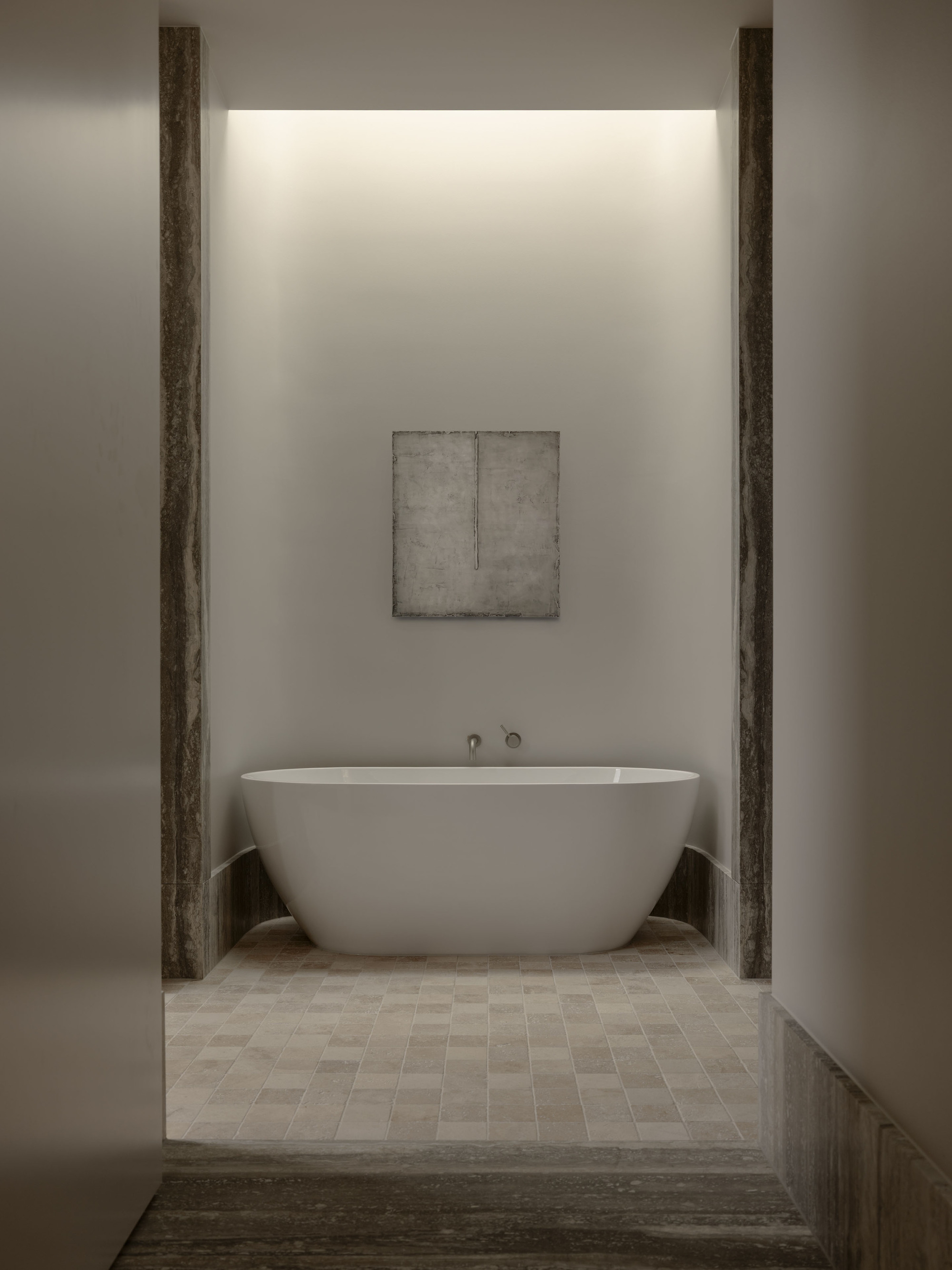 Freestanding white bathtub in an alcove framed by travertine jambs, with a concealed cove light glowing above a small framed artwork