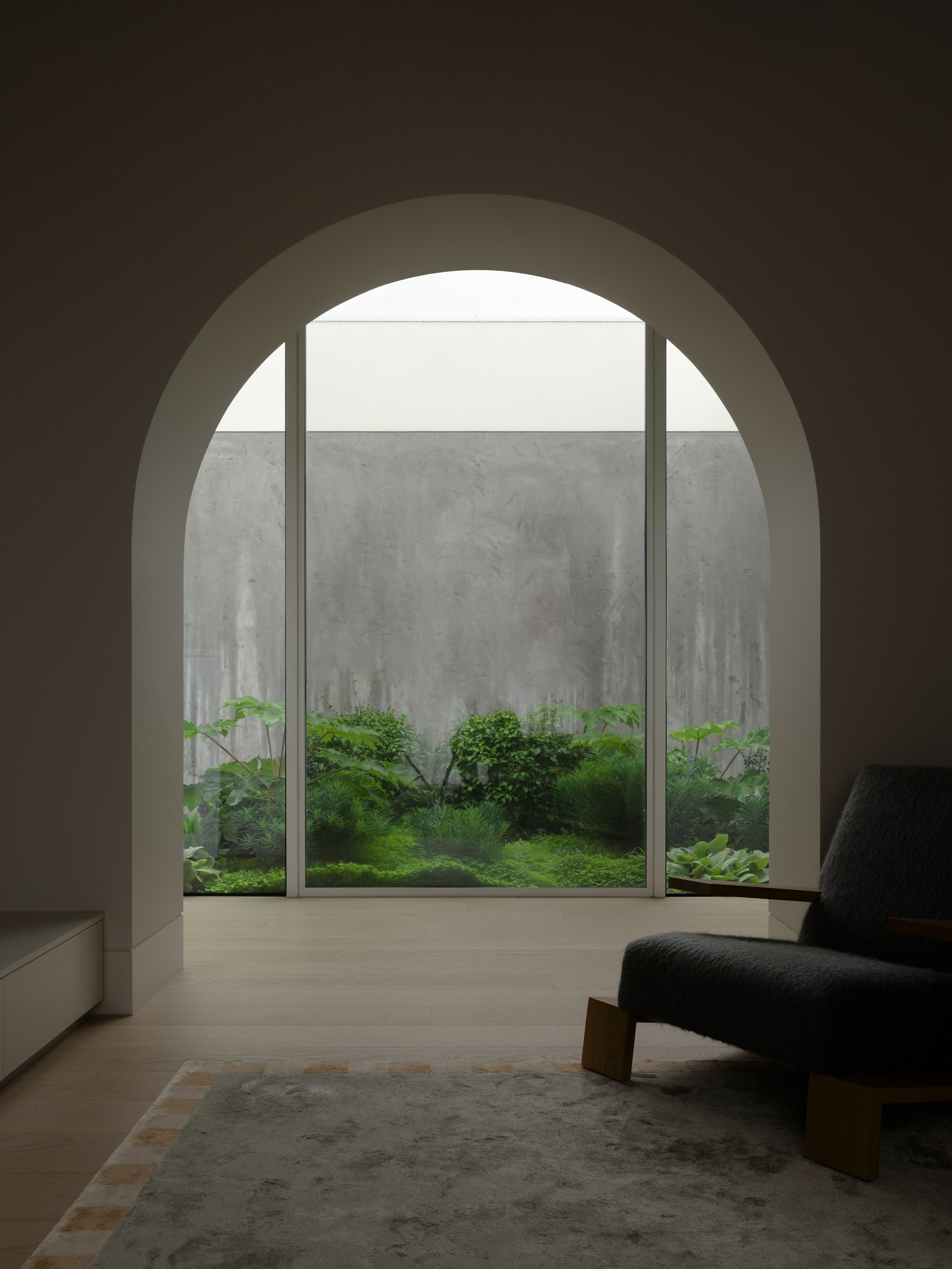 Arched opening framing a glazed courtyard of ferns and hostas against a board-marked concrete wall, with a boucle armchair in the foreground