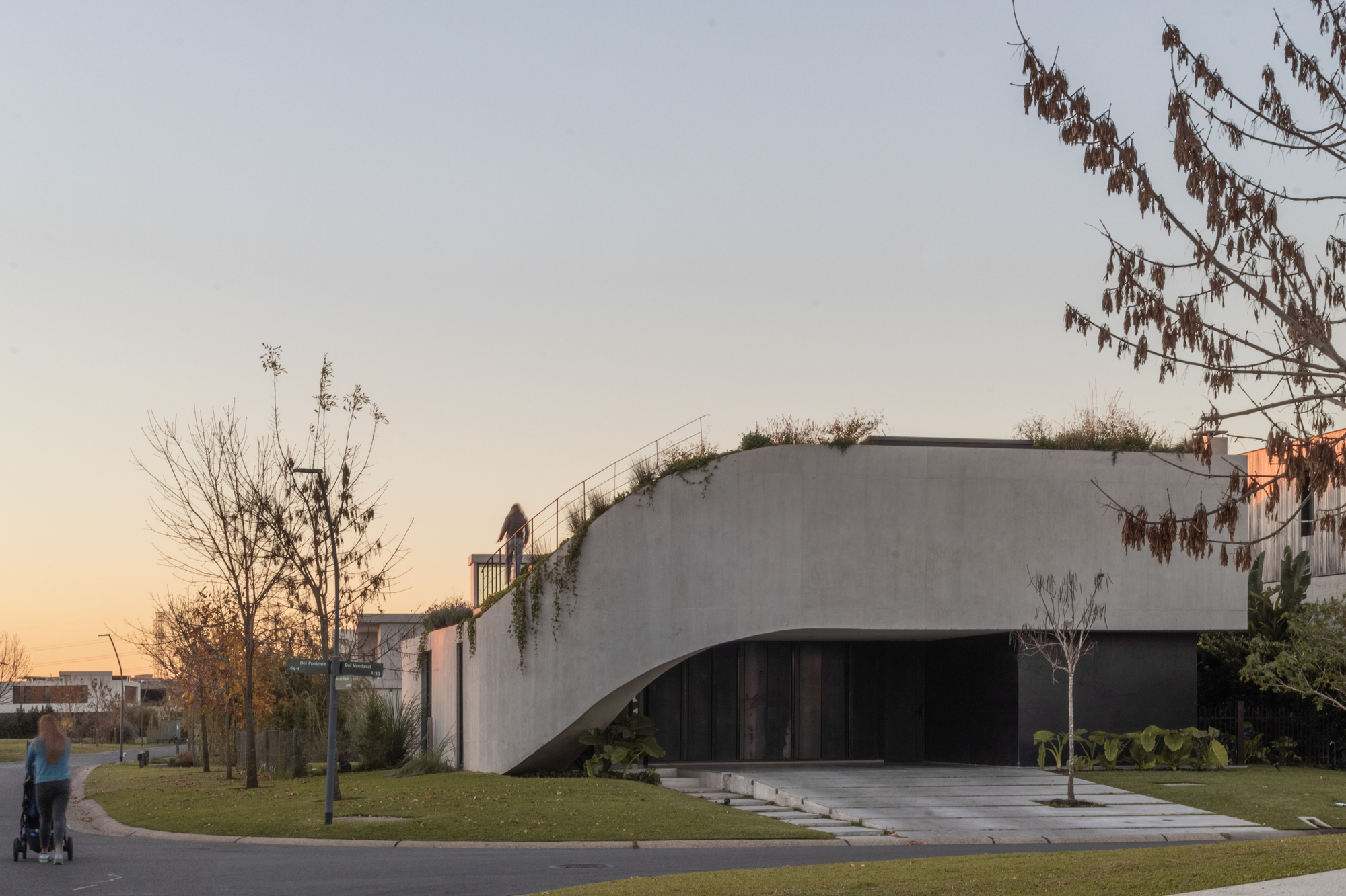 Street corner view of the concrete wall curving over a dark timber garage opening, with a woman pushing a stroller at sunset