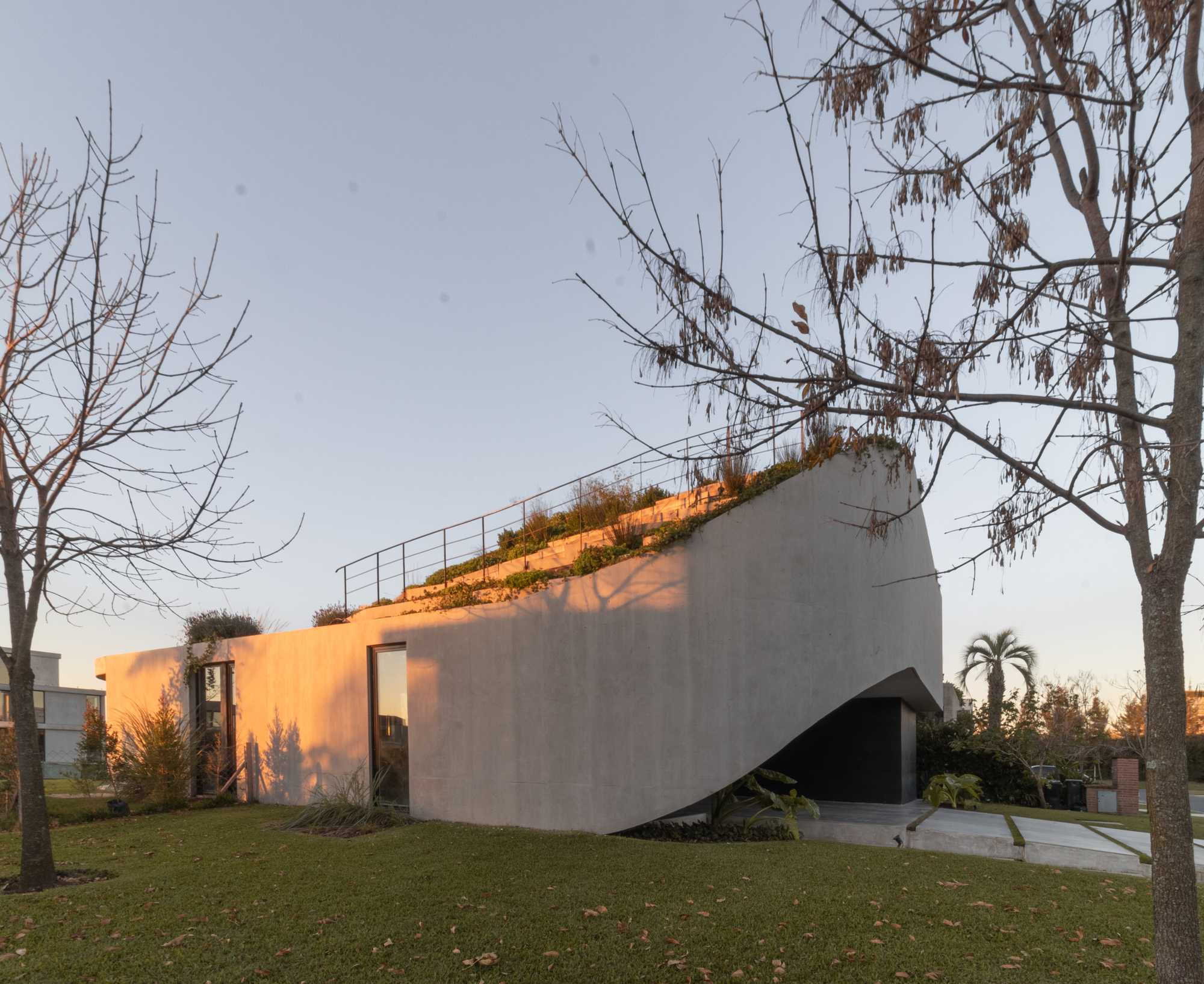 Sloping concrete facade with a green roof edge and steel railing, lit orange by low evening sun