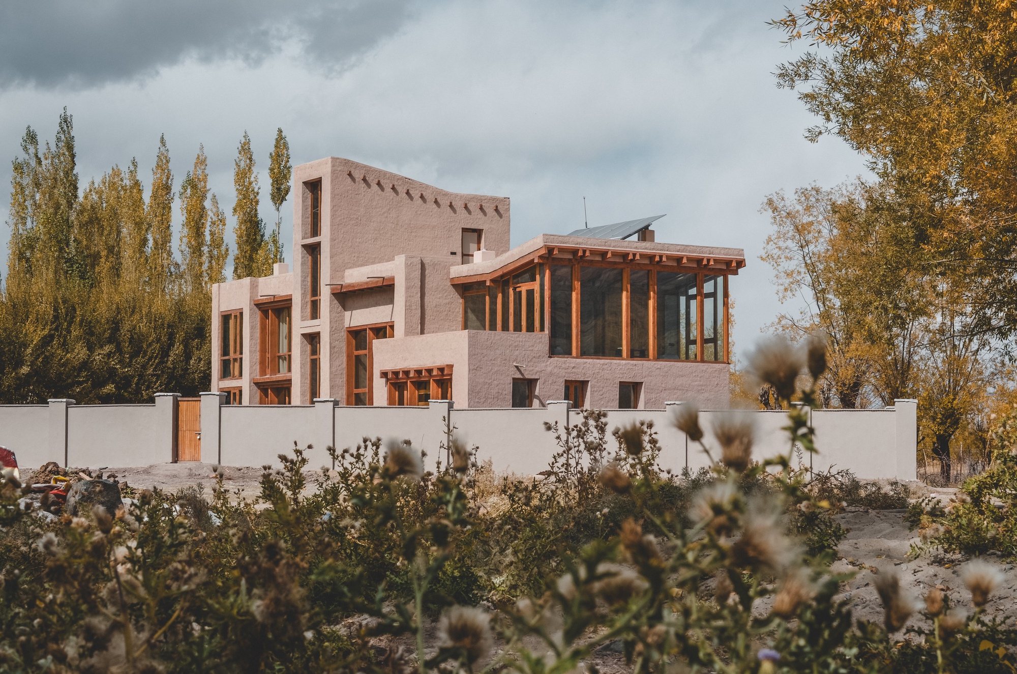 Earth-toned house with timber-framed glazing rising behind a low plaster wall and dry thistles under heavy grey clouds