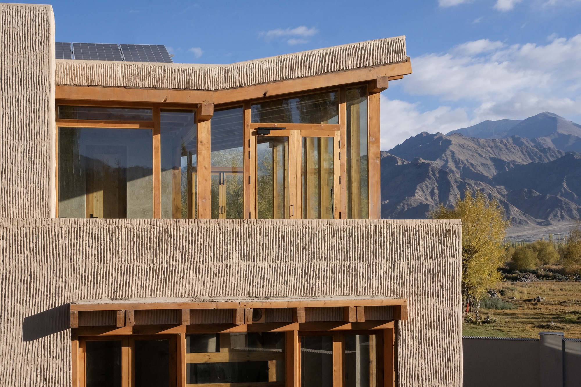 Corner of the textured rammed earth walls with timber-framed glazing and rooftop solar panels against a blue sky and mountain
