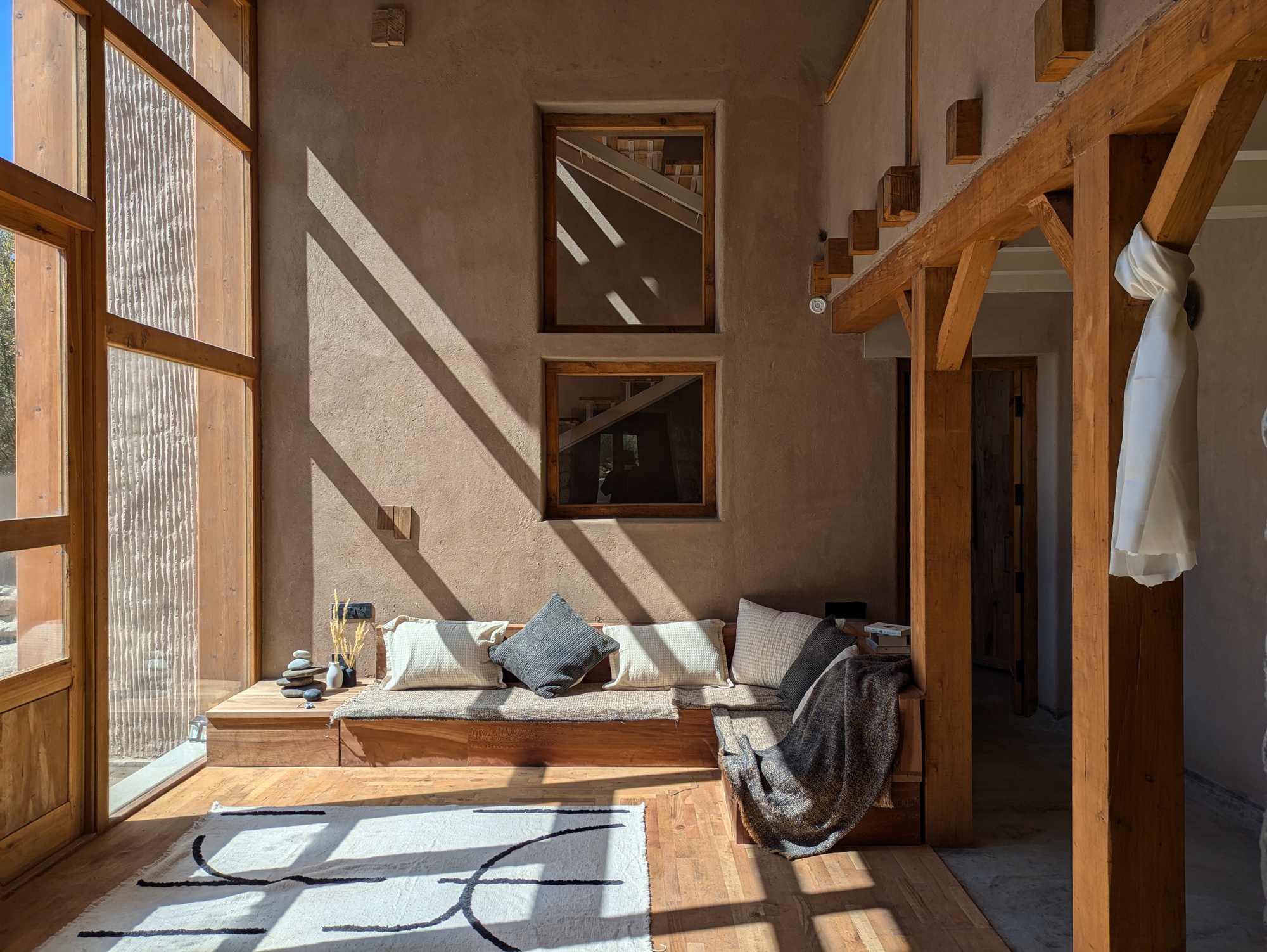 Living corner with built-in daybed, two small square windows and sharp diagonal sunlight falling across the earthen plaster wall