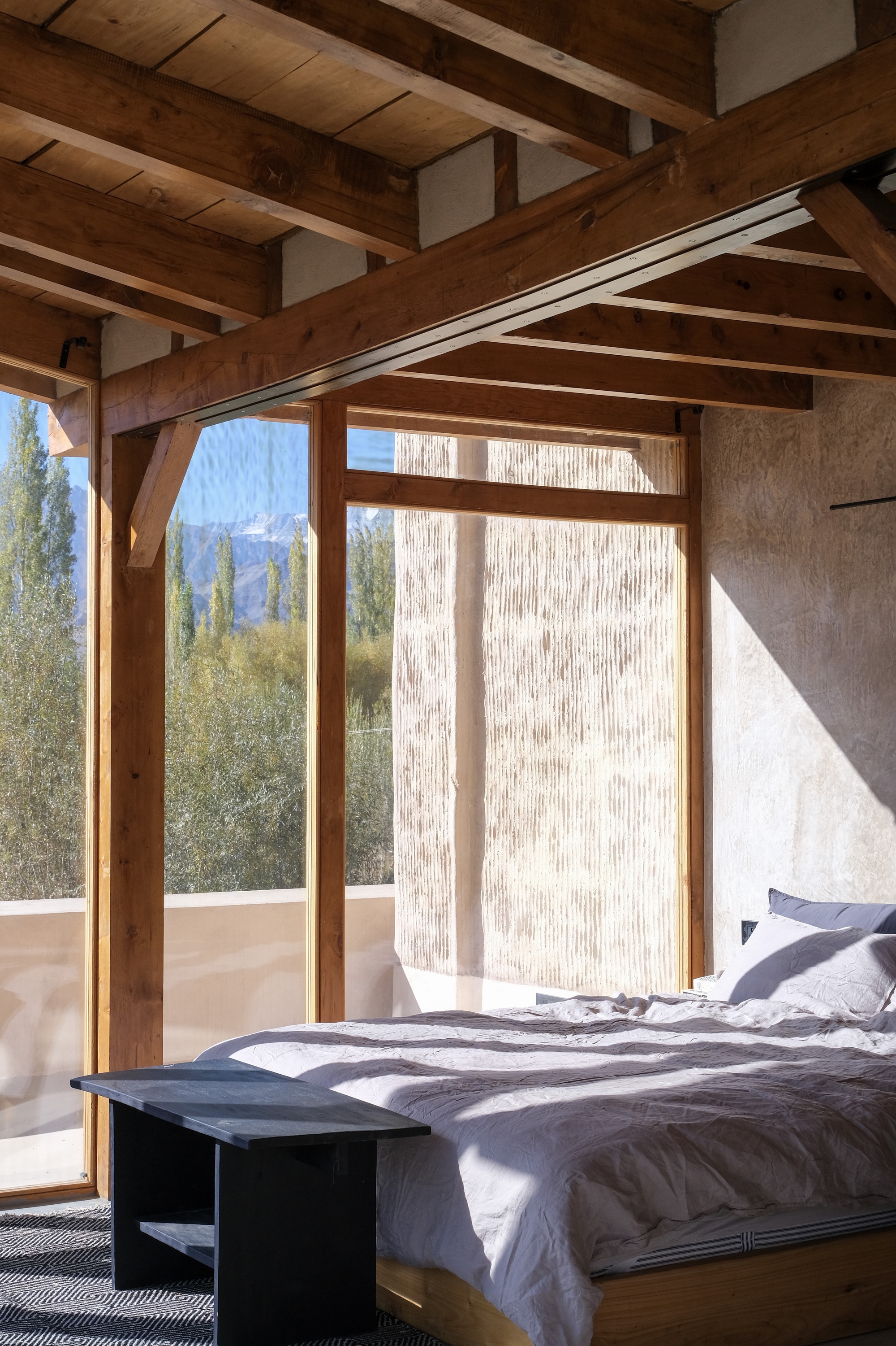 Bedroom under exposed timber beams with a floor-to-ceiling window framing poplars and distant snow peaks in morning light