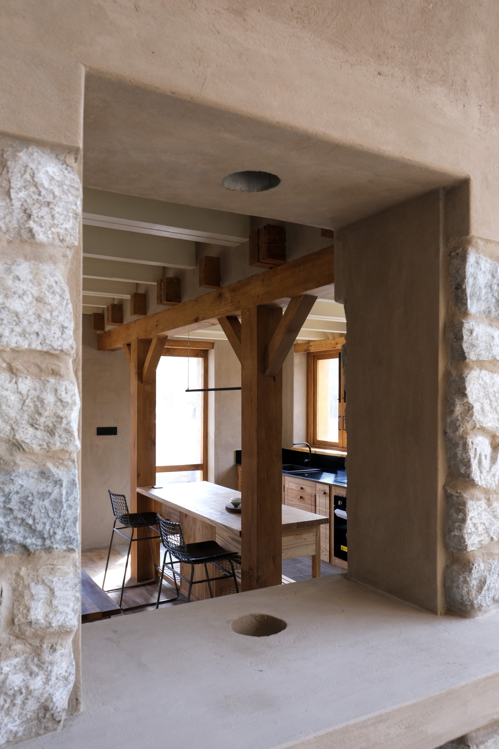 View through a thick plastered wall opening framed by rough stone, toward a timber post and kitchen counter with stools