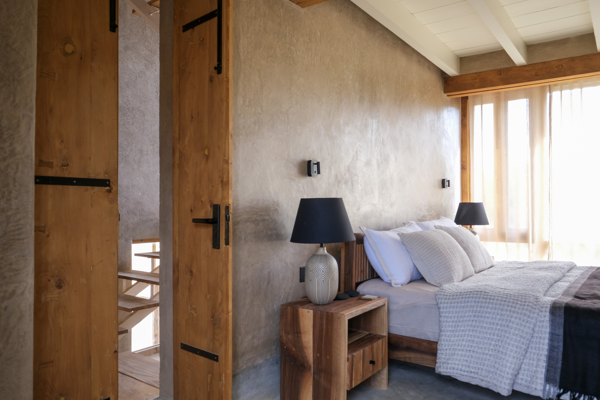 Bedroom with earth-plastered wall, plank doors with black iron hinges and a walnut nightstand beside sheer curtains in afternoon light