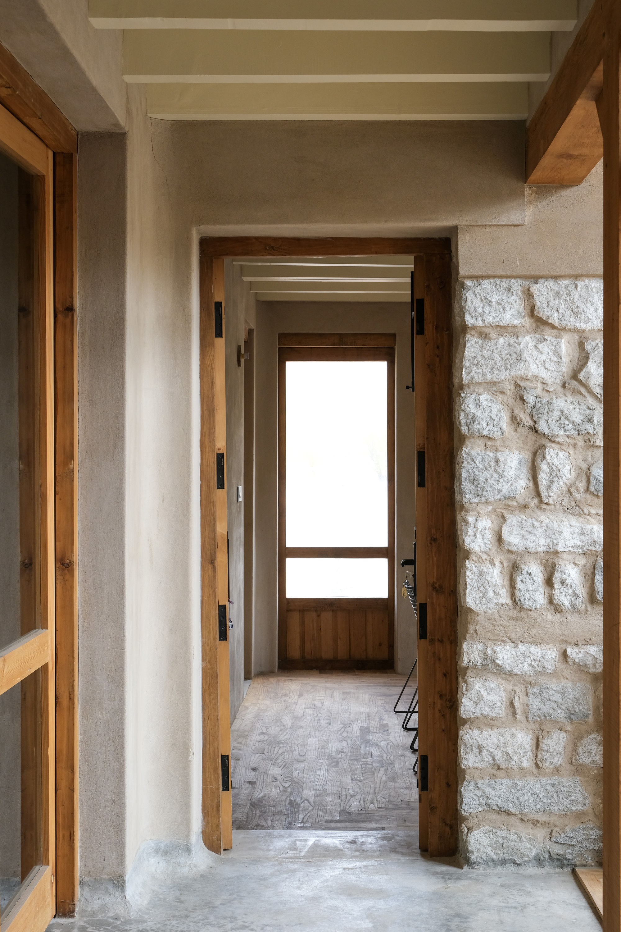 Corridor framed by timber door frames and a granite rubble wall, leading to a glazed door at the far end