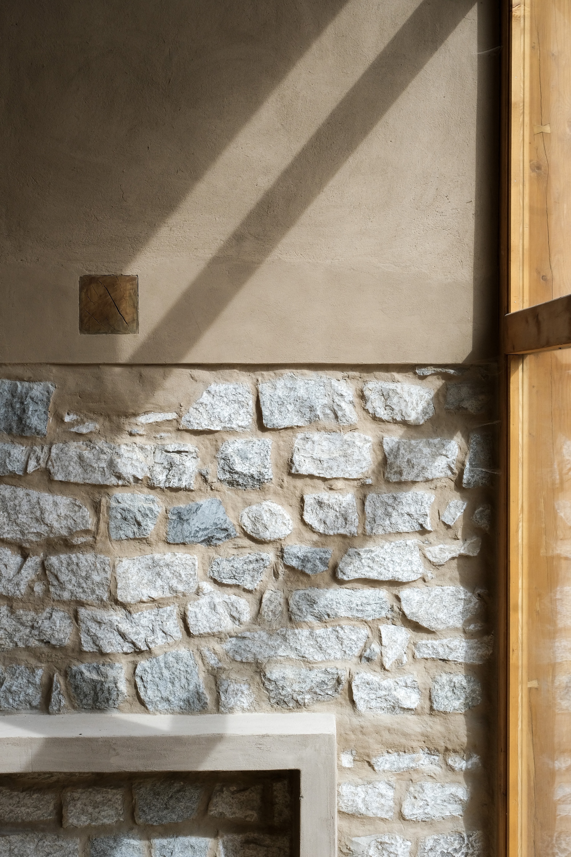 Close-up of rubble stone wall below a clay plaster band, crossed by diagonal shadows from a timber window frame