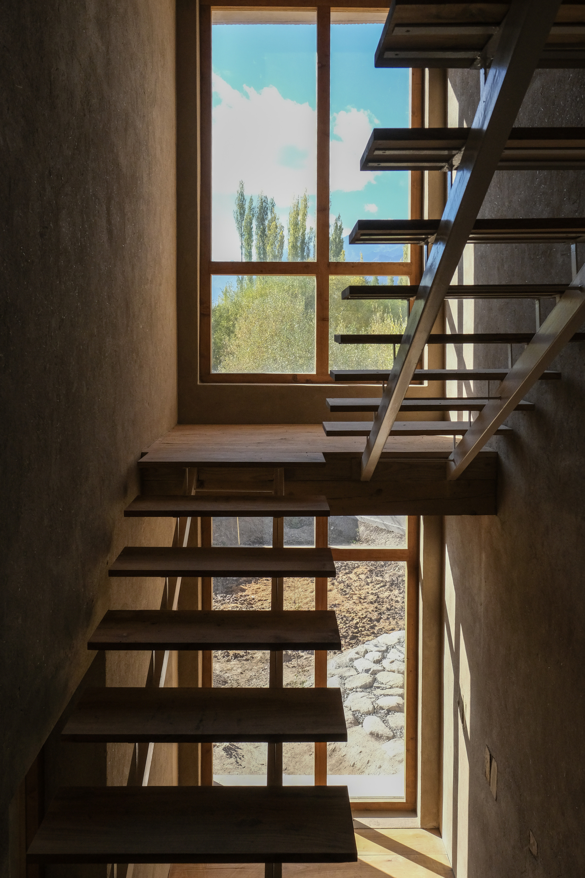 Open-tread timber stair turning at a landing beside a tall window framing poplar trees and a blue sky