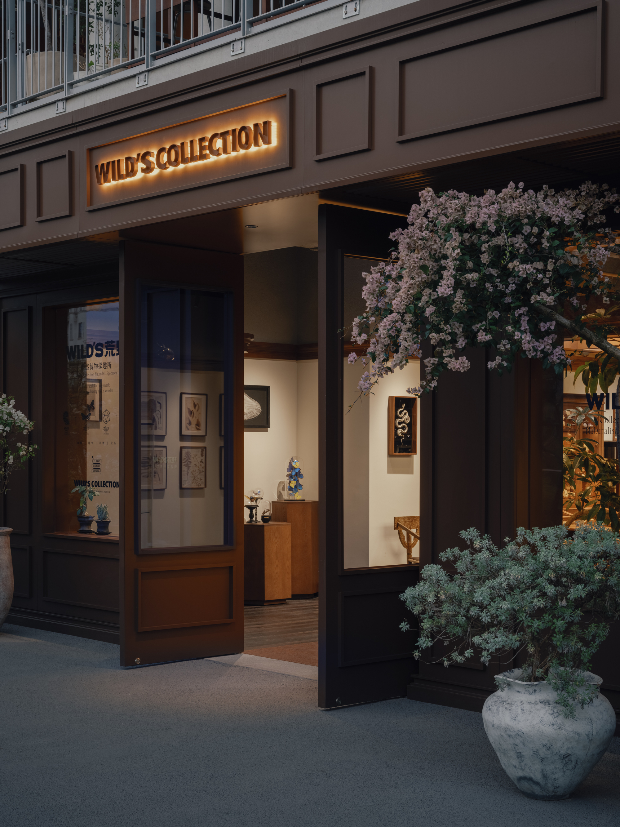 Open timber doors and a glowing wall sign at the shop entrance, with a flowering vine and potted shrub in the evening