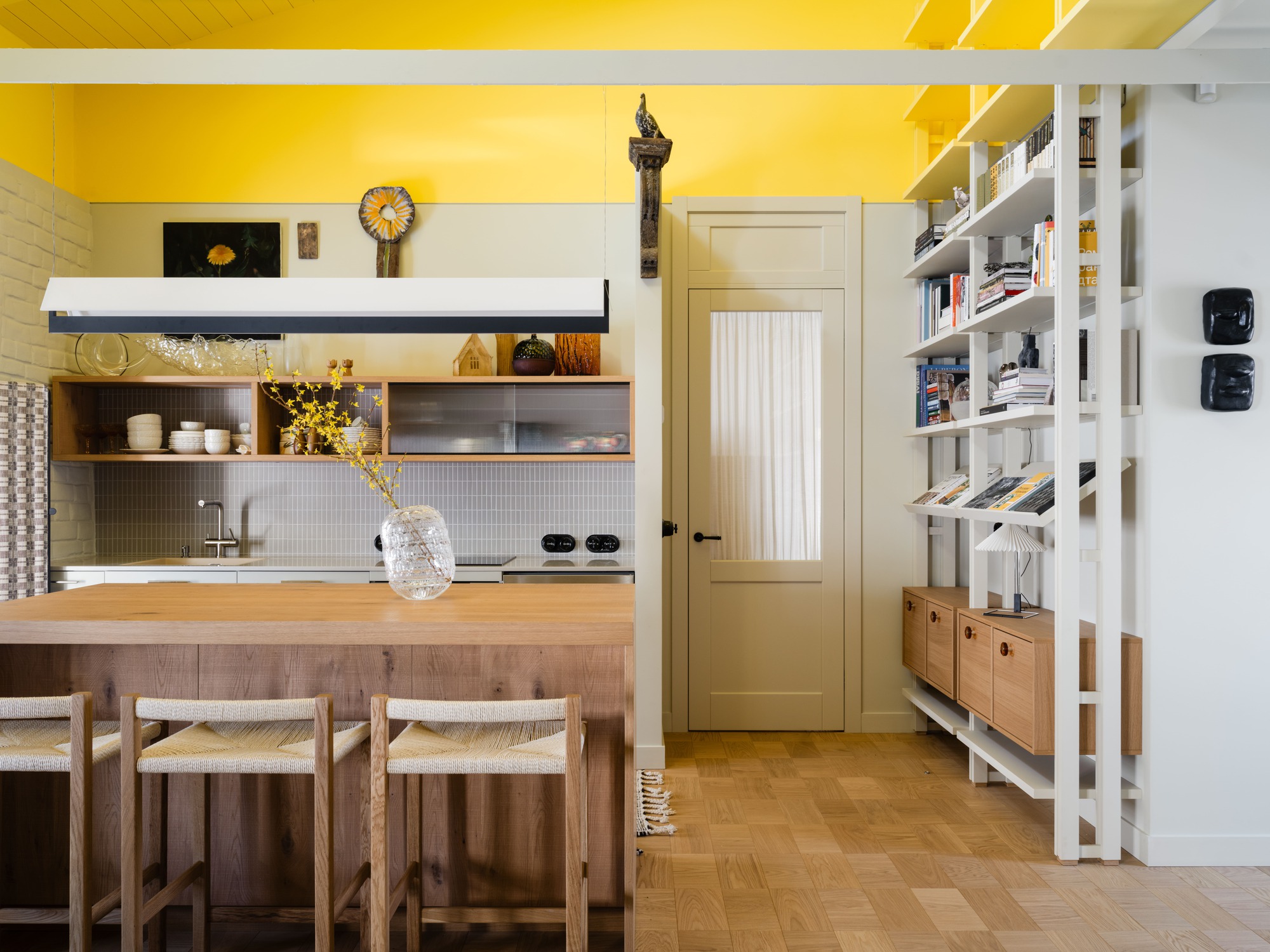 Oak kitchen island with woven-seat stools beside a pale door and white shelving, under a bright yellow upper wall
