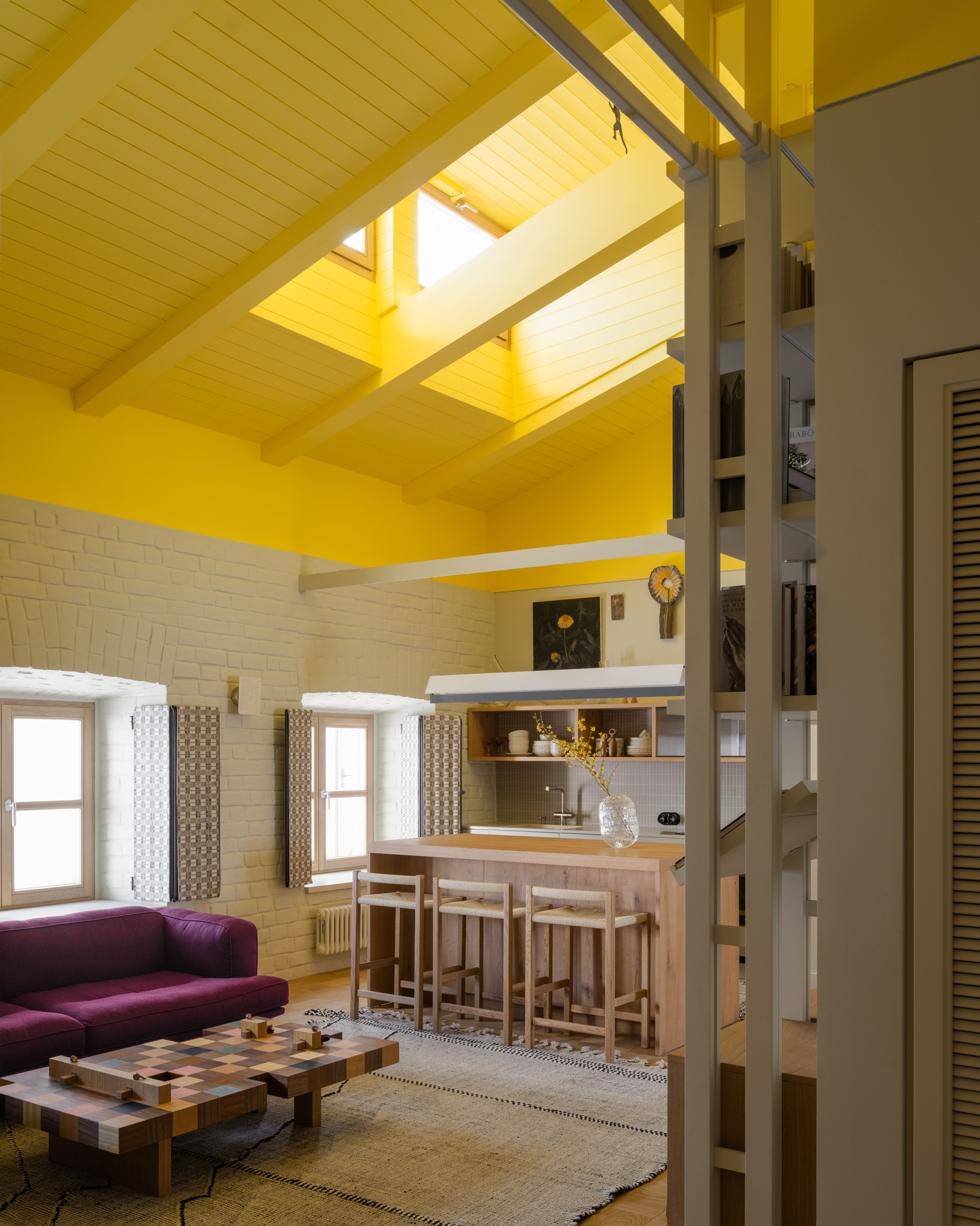 Open living space with yellow-painted beams and skylights above a plum sofa, patterned coffee table and kitchen island in daylight