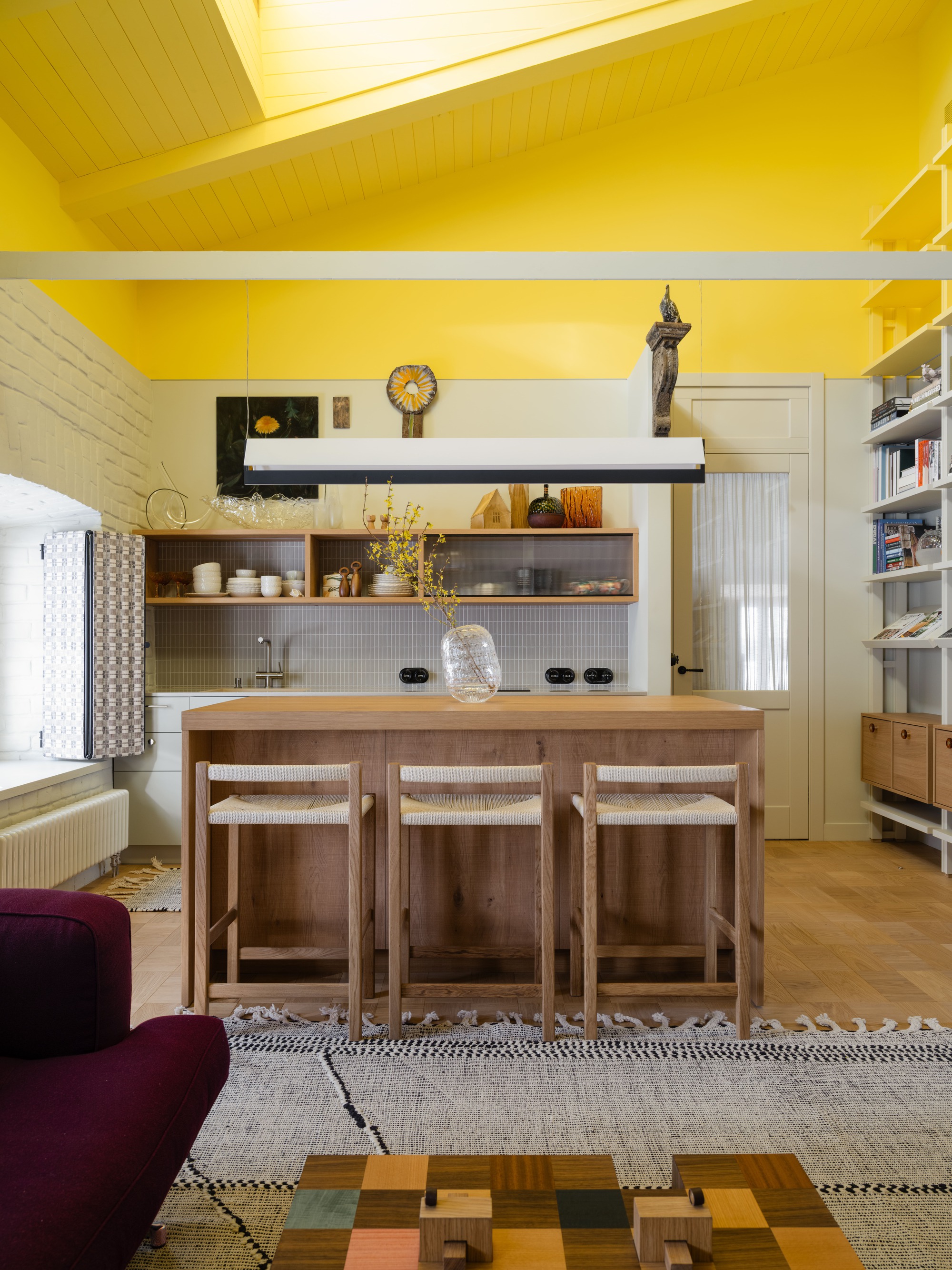Straight-on view of the oak island and three rush-seat stools beneath a sloped yellow ceiling and linear pendant light