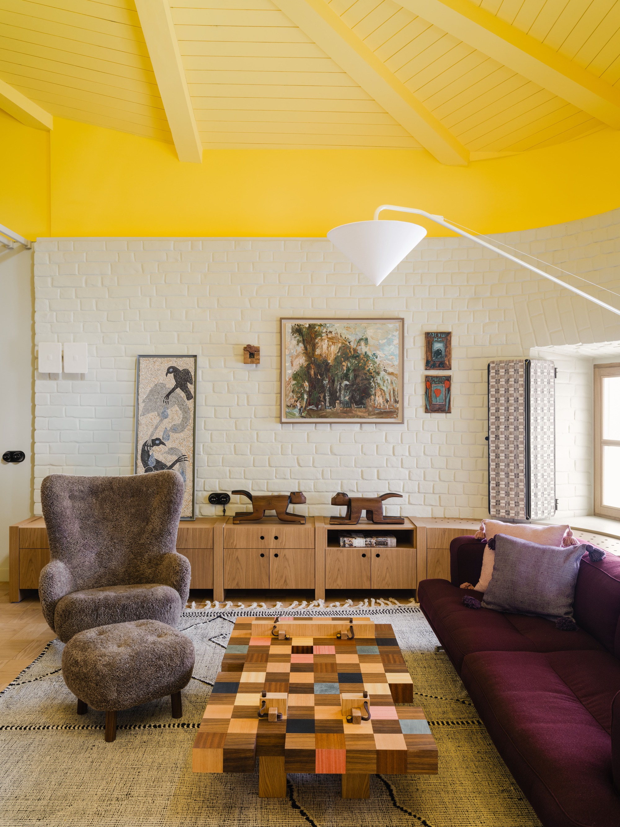 Sitting area with a shaggy armchair, checkerboard wood coffee table and plum sofa against a white painted brick wall
