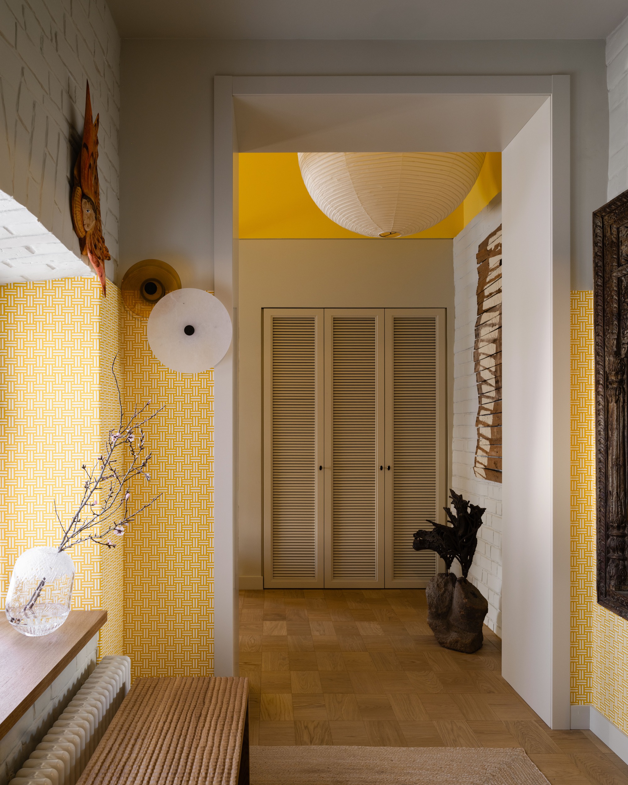 Hallway framed by a white doorway, with yellow patterned wallpaper, a paper globe lamp and louvered cupboard doors at the end