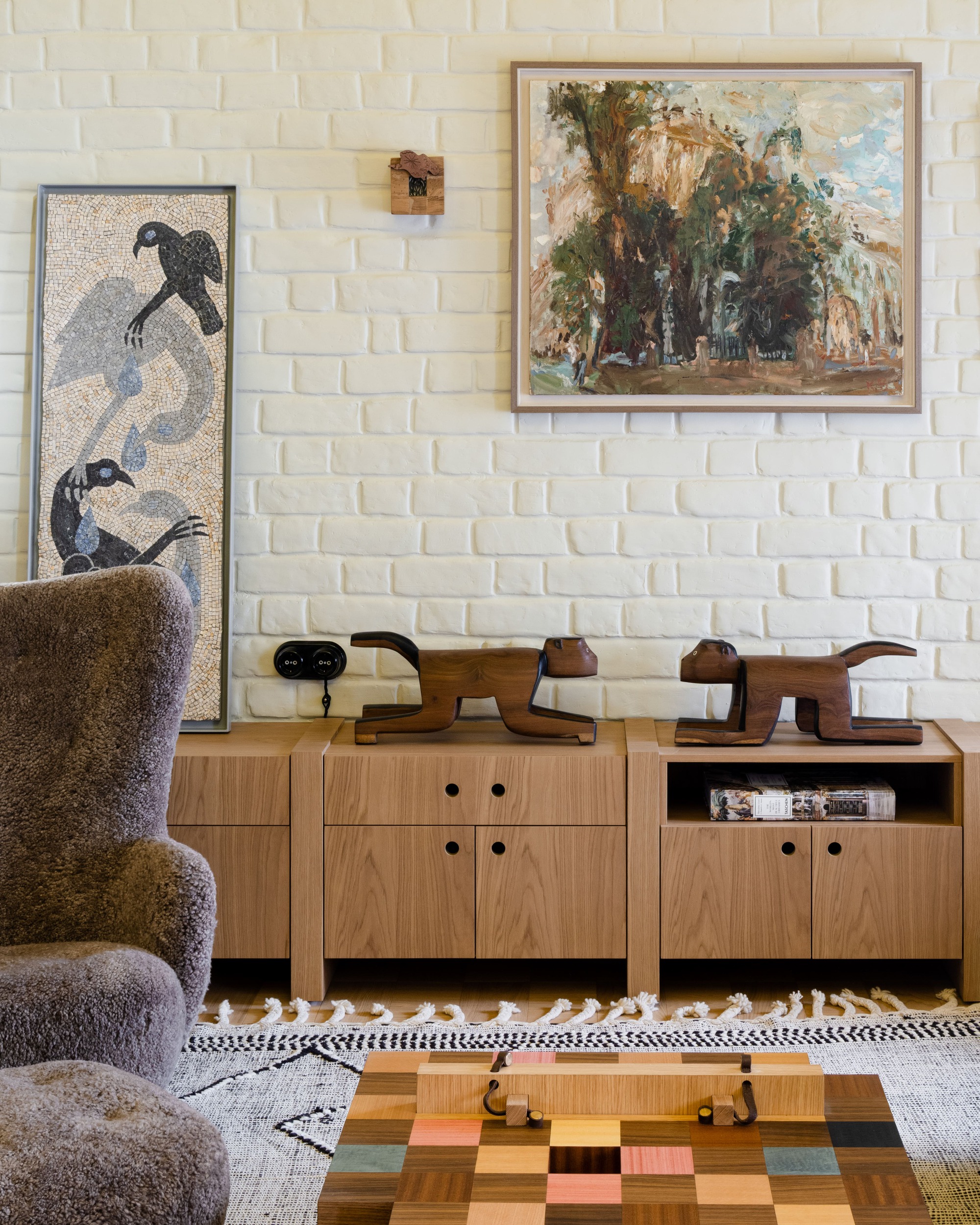 Oak sideboard beneath a white painted brick wall, with two carved wooden animal figures, a mosaic panel and a framed landscape painting