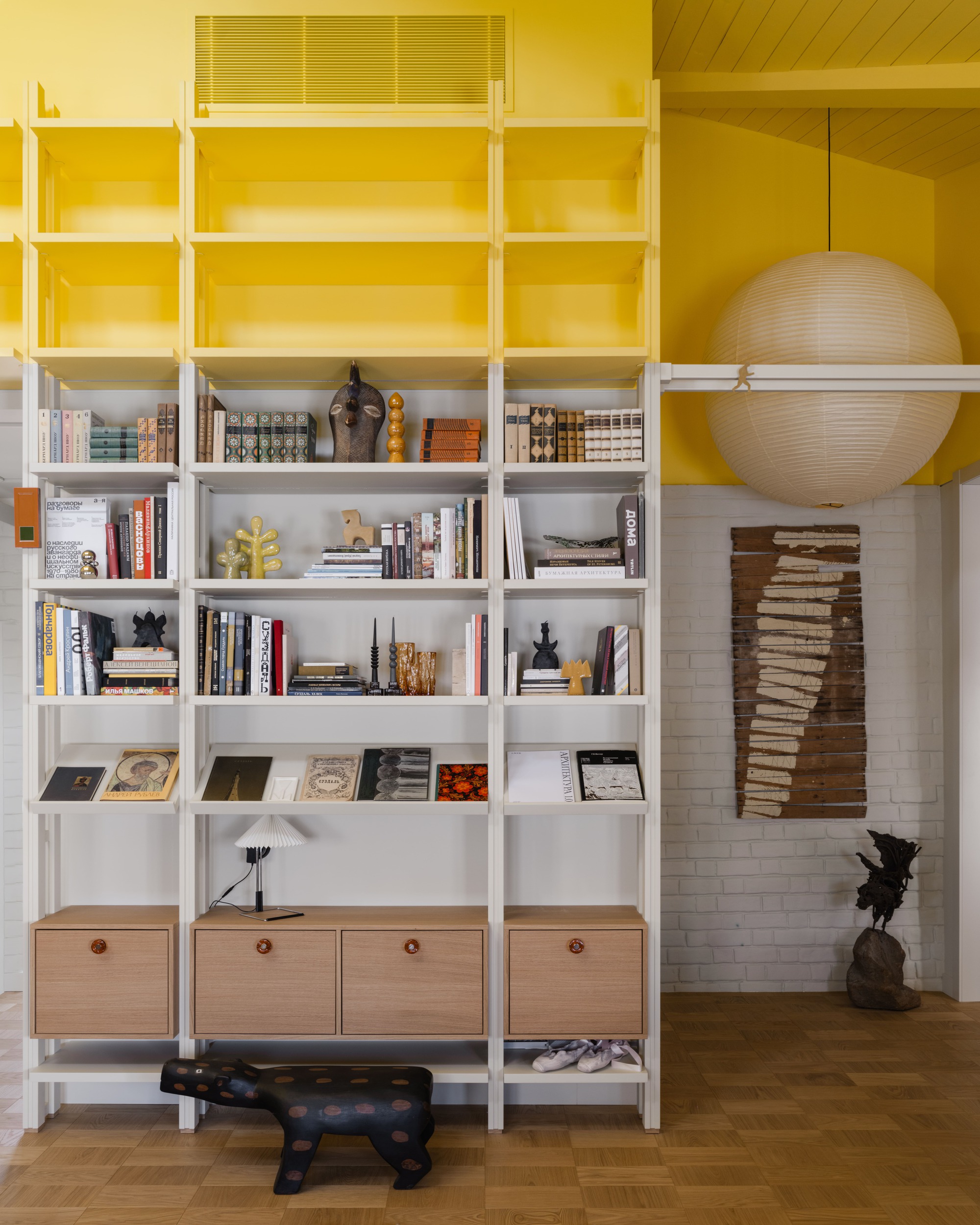 Tall white shelving unit against a yellow wall, with oak cabinet doors, books, objects and a paper lantern hanging beside it