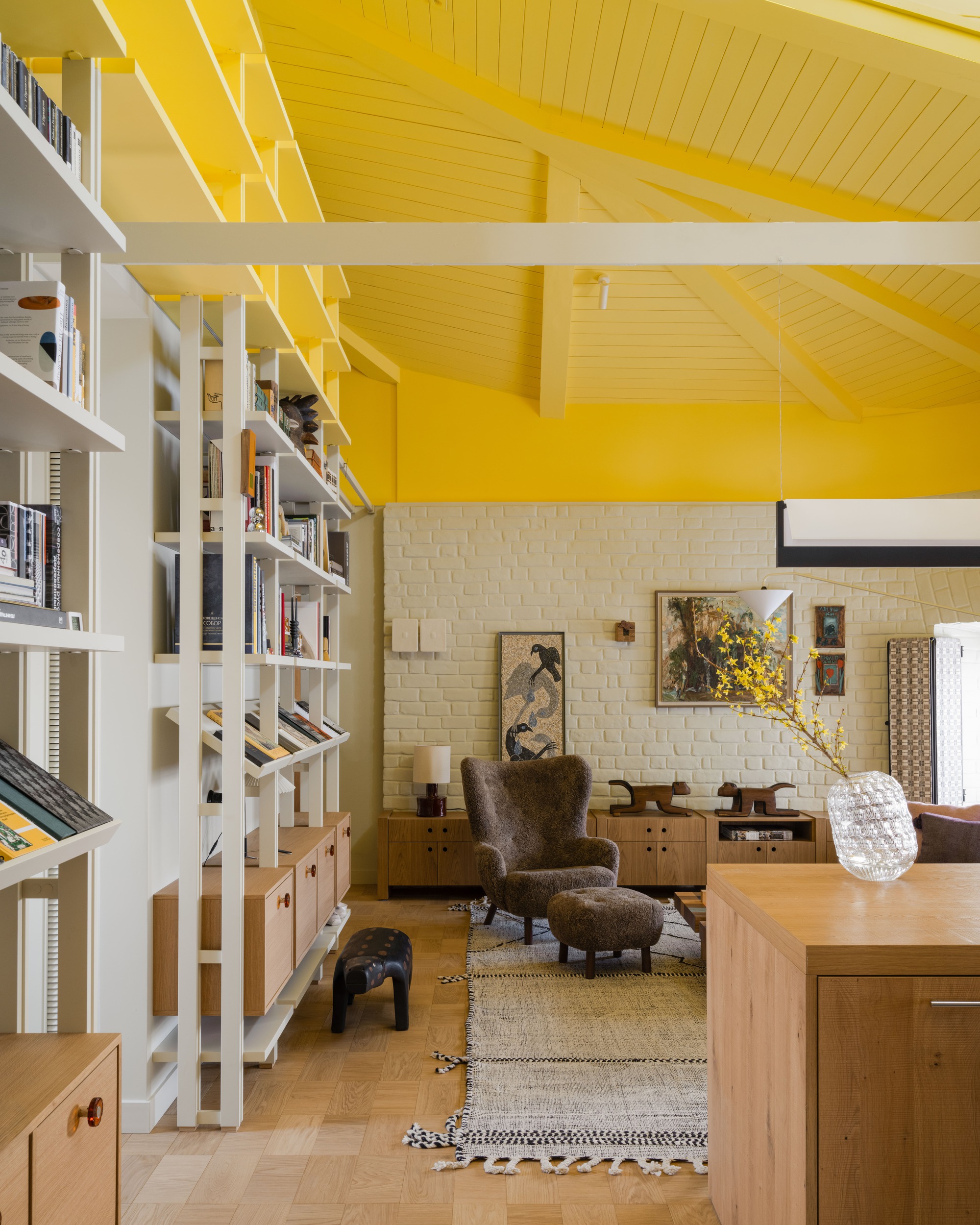Living area seen past bookshelves, with yellow timber ceiling, shaggy armchair, woven rug and a vase of yellow blossom in daylight